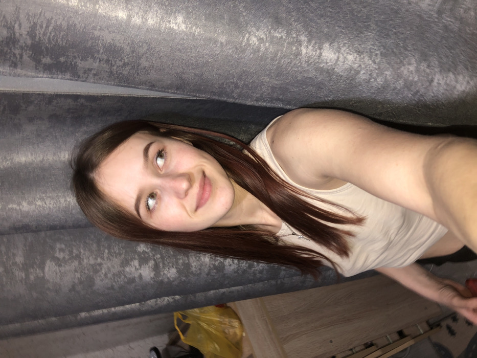 A young adult female is depicted in a close-up selfie taken indoors. She has long, straight, reddish-brown hair, fair skin, and light-colored eyes, with a subtle smile directed towards the right side of the frame. She is wearing a light beige sleeveless top and a thin silver-colored necklace with a small pendant. Her left arm is extended, holding the camera, positioning her right shoulder against a dark grey, vertically folded, textured curtain that dominates the background behind her. To the left of the subject, a light-colored wooden furniture item is partially visible, topped by a yellow plastic bag and a dark, indistinct object. The lighting appears to be artificial. The specific city of Foki, Russia, as requested, cannot be corroborated from the visual evidence presented in the image.
