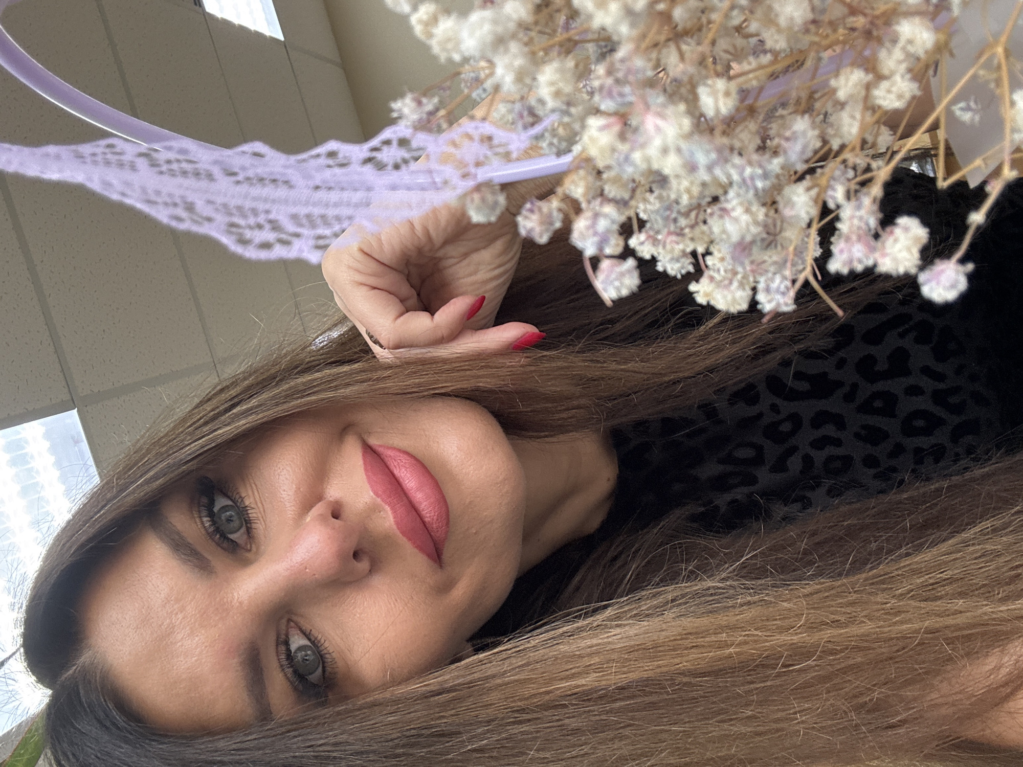 A close-up, selfie-style photograph shows a woman with long, brown hair, blue eyes, and pink lipstick. She is looking directly at the camera with a slight smile. Her right hand is raised, resting on a purple basket handle, and she is holding a bunch of small, white flowers. The woman is wearing a black top with a leopard print pattern. The background appears to be an indoor office environment with fluorescent lighting and a white ceiling. The location is Chaikovsky, Russia.