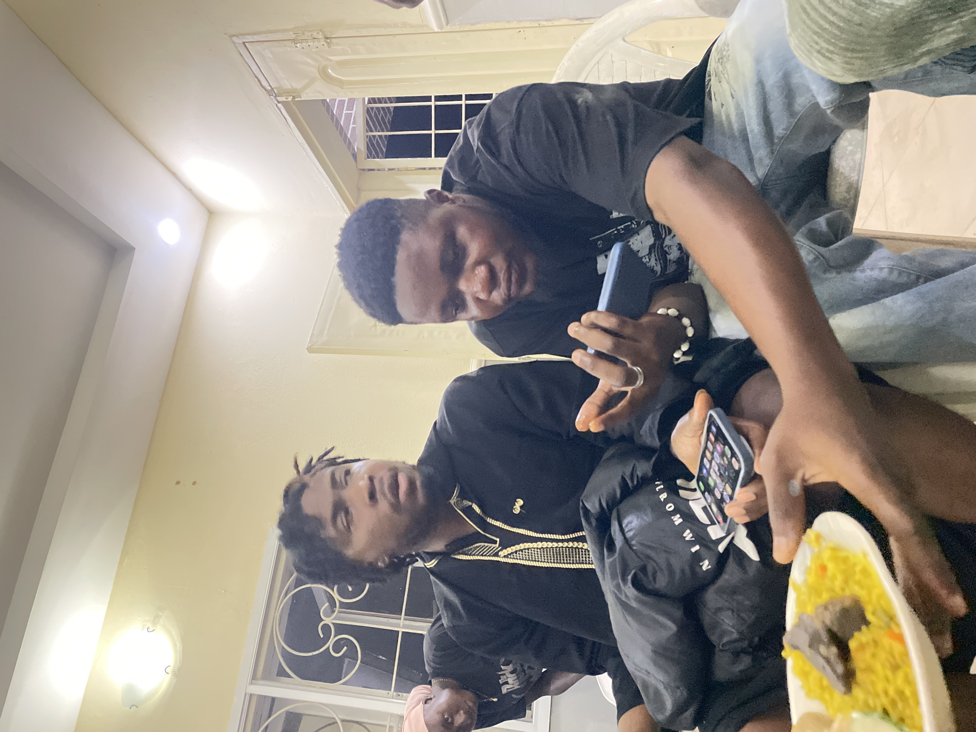 Two dark-skinned men are inside a building, possibly in Jos, Nigeria, based on the information given. One man, wearing a black shirt, sits in a white chair, looking at his phone. The other man, also dark-skinned and wearing a black outfit with gold trim, sits lower, holding a phone and a plate of yellow rice with meat. Another person can be seen partially in the bottom left of the photo. A window with white bars and white walls can be seen in the background, along with a ceiling with recessed lighting.