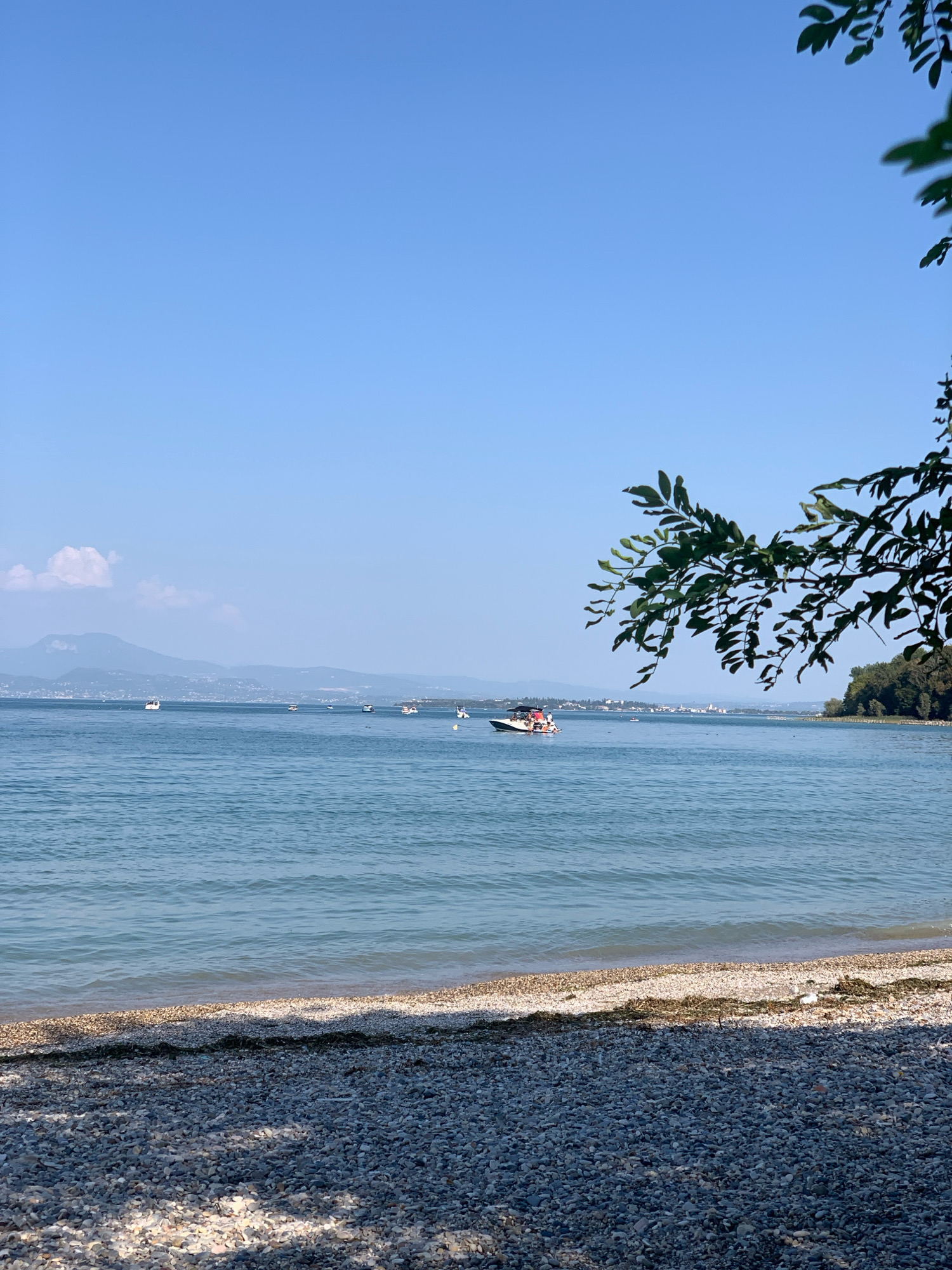 The image depicts a serene waterscape and a pebble beach in Padenghe sul Garda, Italy. The clear blue sky stretches across the top of the frame, meeting the tranquil blue waters of a lake. In the distance, hazy mountains form a picturesque backdrop, with a few white clouds adding texture to the sky. Several small boats are scattered across the water, one of which appears to be a speed boat with people on board. The foreground is dominated by a pebble beach, composed of a multitude of small, gray, and white stones, with some dried seaweed and debris along the water's edge. Lush green foliage from a tree branch hangs into the frame from the upper right corner, casting dappled shadows on the beach. The overall impression is one of a peaceful and sunny day by the water.