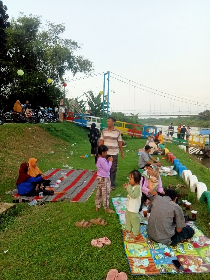 The scene is a grassy park in Jakenan, Indonesia (coordinates: -6.758, 111.075). A group of people are gathered under a suspension bridge. Several people are sitting on picnic blankets. There are two children in pink dresses, one is standing and looking at the camera, the other is sitting on a blanket looking down. There are several people wearing headscarves, some sitting, some standing and talking. An older man with a striped shirt is standing, looking at the camera. There are several motorcycles parked on the grass next to a dirt path. The bridge has colorful wooden handrails. The ground is covered in green grass. Several plastic cups are visible on the picnic blankets. The sky is bright blue, with a few white clouds. The trees on the other side of the bridge are green. A man is sitting on a colorful patterned blanket, looking down at a cell phone. Several people are walking on the bridge in the distance. There are a few pieces of trash on the ground.