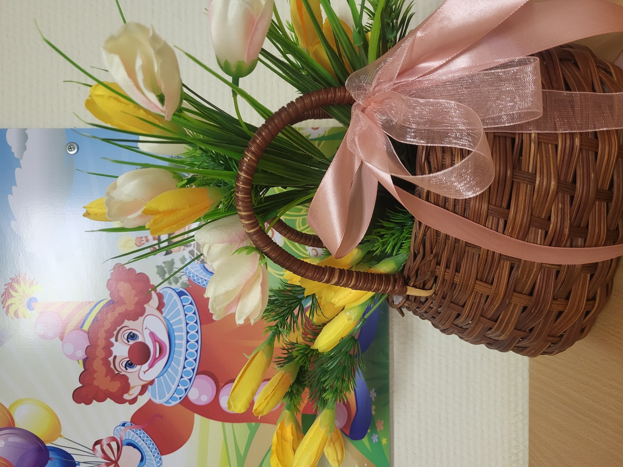 The image shows a brown wicker basket filled with artificial white and yellow tulips and green foliage. A pink ribbon is tied around the basket's handle. The basket is hanging on a wall next to a picture featuring a clown with balloons. The image was taken in Chaikovsky, Russia.