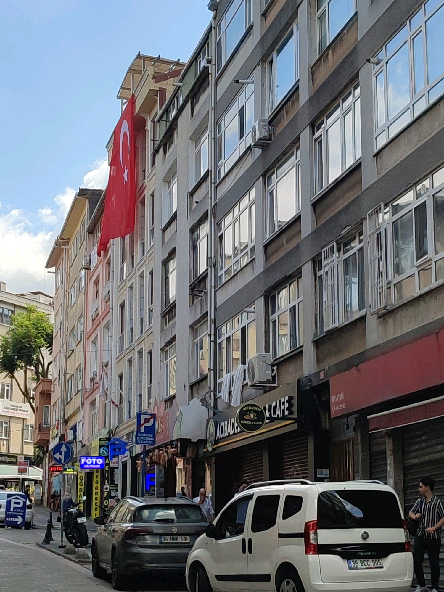The image shows a street scene in Kadıköy, Turkey, on a clear day with scattered clouds. The foreground features a white Fiat Fiorino minivan parked on the right, with its rear end visible. A person in a striped shirt stands to the right of the minivan, looking towards the left. To the left of the minivan, a dark grey hatchback car is parked. Behind the cars, a row of multi-story buildings lines the street, displaying a mix of architectural styles. A large Turkish flag is prominently displayed hanging from one of the buildings. Several businesses with signage are visible on the ground floor of the buildings, including "ACIBADEM POKER & CAFE" and "FOTOĞRAF". Traffic signs, including parking signs, are also present. The street appears to be paved, with a sidewalk running alongside the buildings. The overall impression is that of a typical urban street in a bustling commercial district.