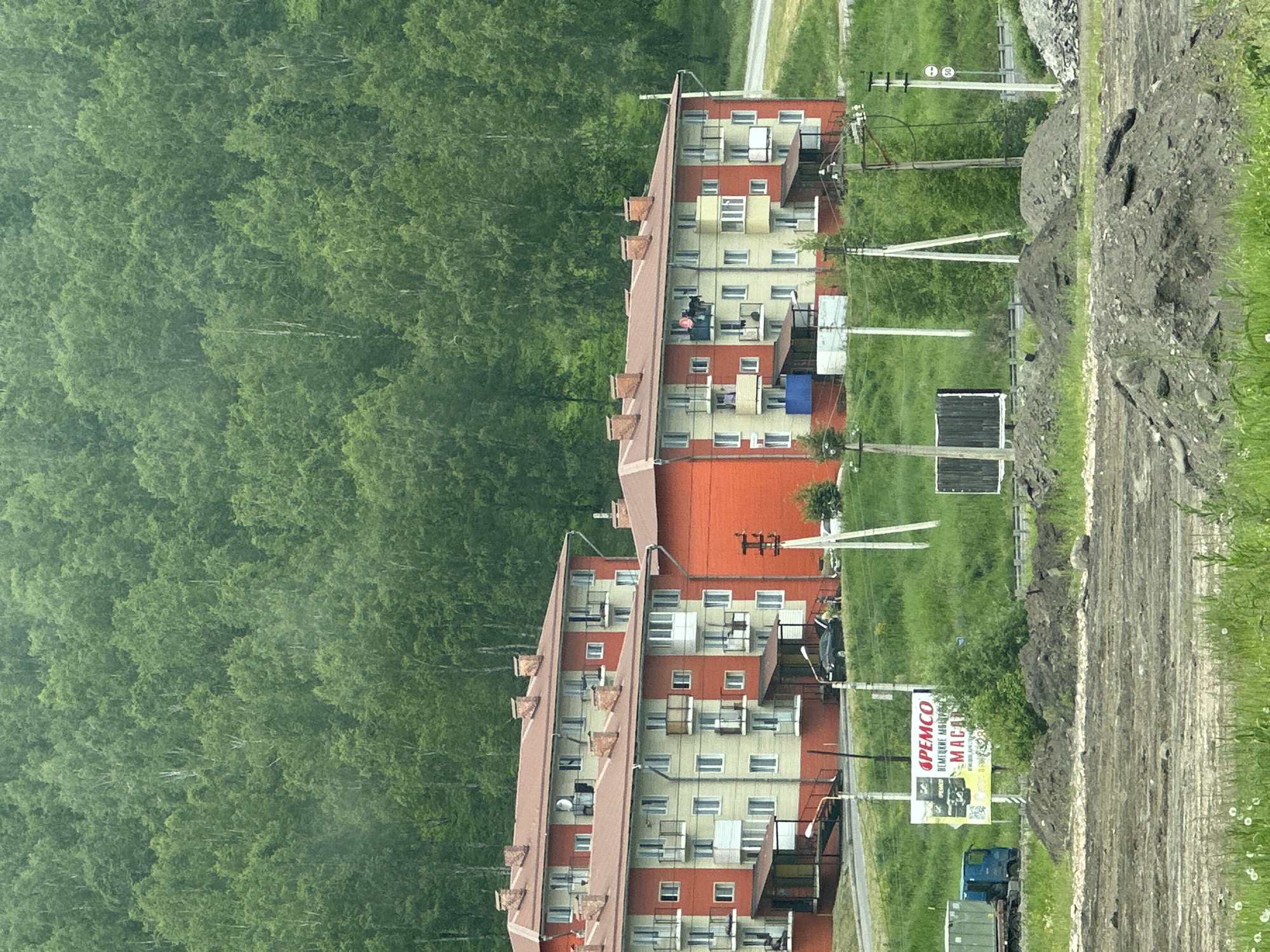 The image depicts a residential area in Slyudyanka, Russia, with multi-story buildings set against a backdrop of a dense forest. Two apartment buildings, painted in a combination of yellow and orange, are prominently featured. These buildings have red-tiled roofs with visible chimneys and multiple balconies with railings. Several windows are visible on the facades of the buildings.
In front of the buildings, a street is visible, with a parked black car and a few people standing near it. A blue truck with a cargo container is parked on the left side of the image. A billboard with Russian text and images is also present, advertising "PEMCO".
The environment consists of grassy slopes, some areas with piles of dirt or gravel, and utility poles with wires. The overall impression is a functional, somewhat utilitarian residential neighborhood bordering a natural wooded area. The image appears to be taken during daylight, with overcast conditions contributing to a muted color palette.