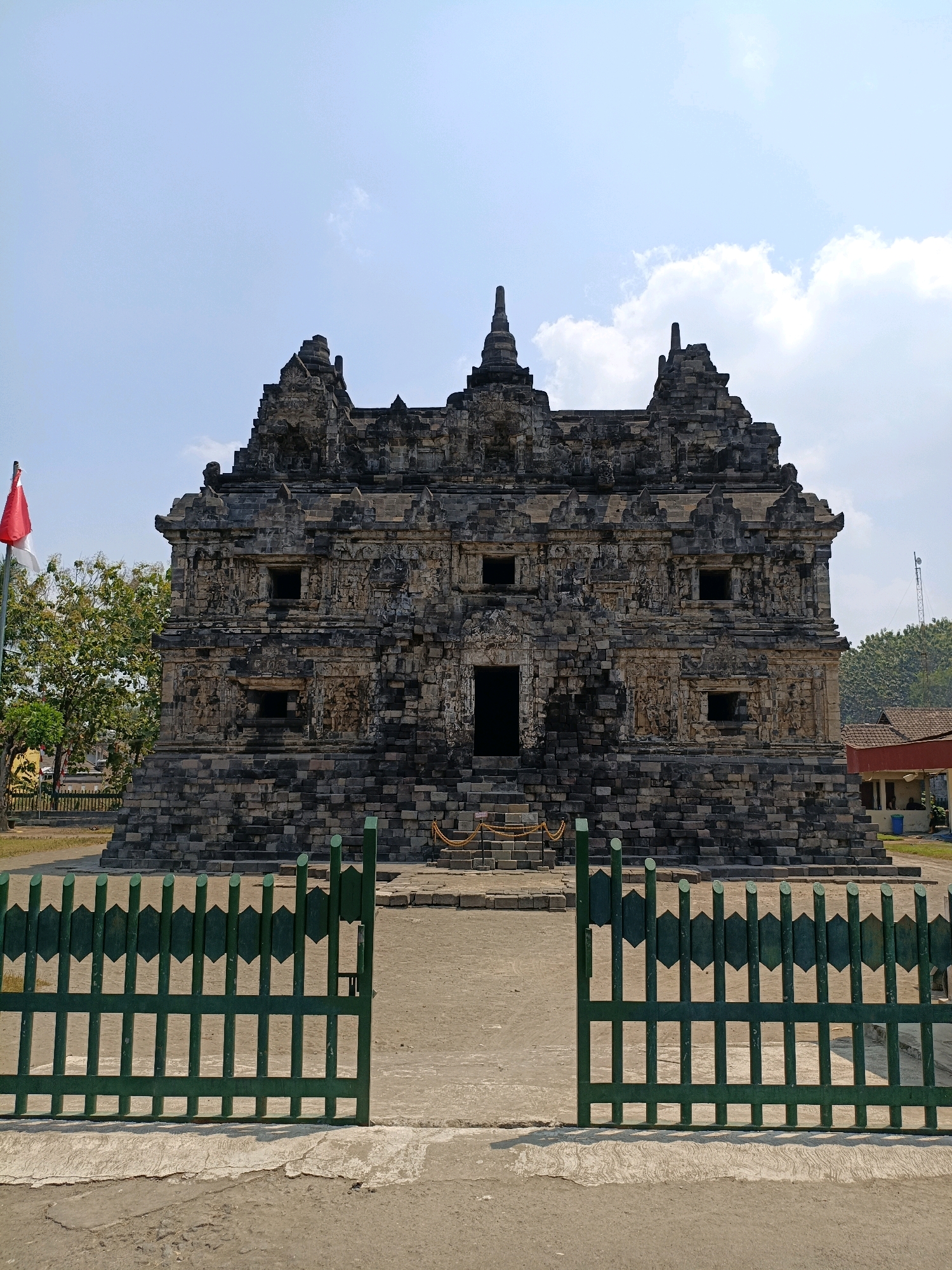 The image shows the Candi Banyunibo, a Hindu temple located in Tirtomartani, Sleman Regency, Yogyakarta Special Region, Indonesia. The temple is constructed from gray stone and features intricate carvings and tiered roofs. A red and white flag is visible to the left of the temple. In the foreground, there is a green metal fence with an open gate. The sky is blue with scattered clouds, and trees are present in the background.