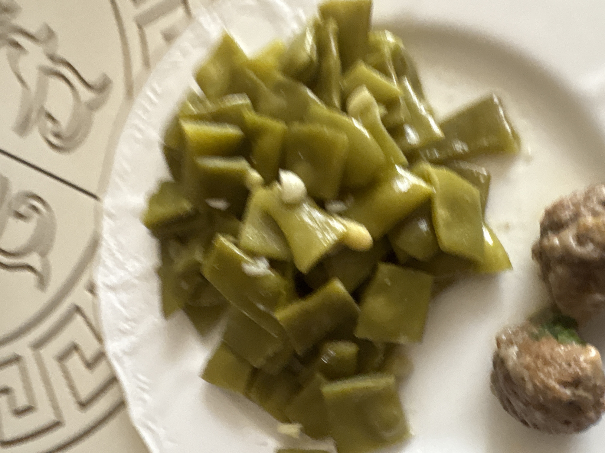 A plate of sliced green beans and two meatballs is on a patterned surface. The green beans are bright green and appear to be cooked, with visible pieces of garlic interspersed. The meatballs are brown and have a textured surface. The plate is white and sits on a surface that has a repeating geometric pattern, possibly a tablecloth or placemat. The background is blurred, but suggests an indoor setting. The image quality is poor, with blurriness and low resolution, making precise identification of all details difficult. The scene is likely a meal prepared at home. There is no information available regarding the location in the city of Georgiyevsk, Russia.