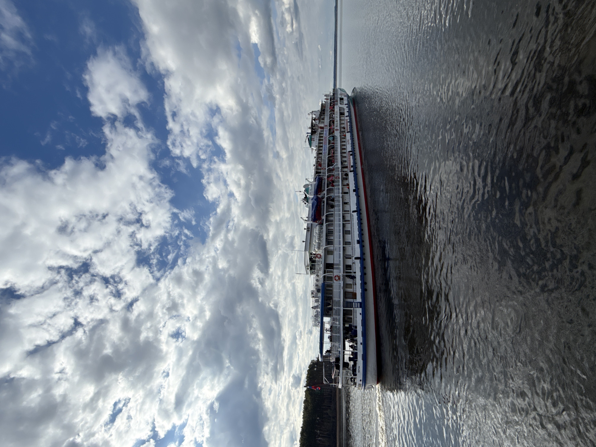 The image captures a large, multi-deck river cruise ship, identified by the visible text "ВАСИЛИЙ ЧАПАЕВ" (Vasiliy Chapayev) and "Лотос Тур" (Lotus Tour) on its hull, sailing on a wide body of water. The ship is primarily white with a red stripe near the waterline and blue detailing on its upper decks and superstructure. Numerous windows and balconies are visible along its length, indicating passenger cabins, and several lifeboats are mounted on the upper levels.
The vessel is positioned on calm water that shows gentle ripples and subtle reflections, suggesting either slow movement or a slight current. In the distance, on the opposite bank, a dark green treeline composed of dense conifers stretches across the horizon. The sky overhead is partly cloudy, featuring patches of bright blue interspersed with large, soft white and grey cumulus clouds. The scene is brightly lit, indicating daytime, likely late morning or afternoon, with fair weather conditions. The overall atmosphere is one of serene travel. This setting is located in or around Chaikovsky, Russia, consistent with the region's large rivers and reservoirs.
