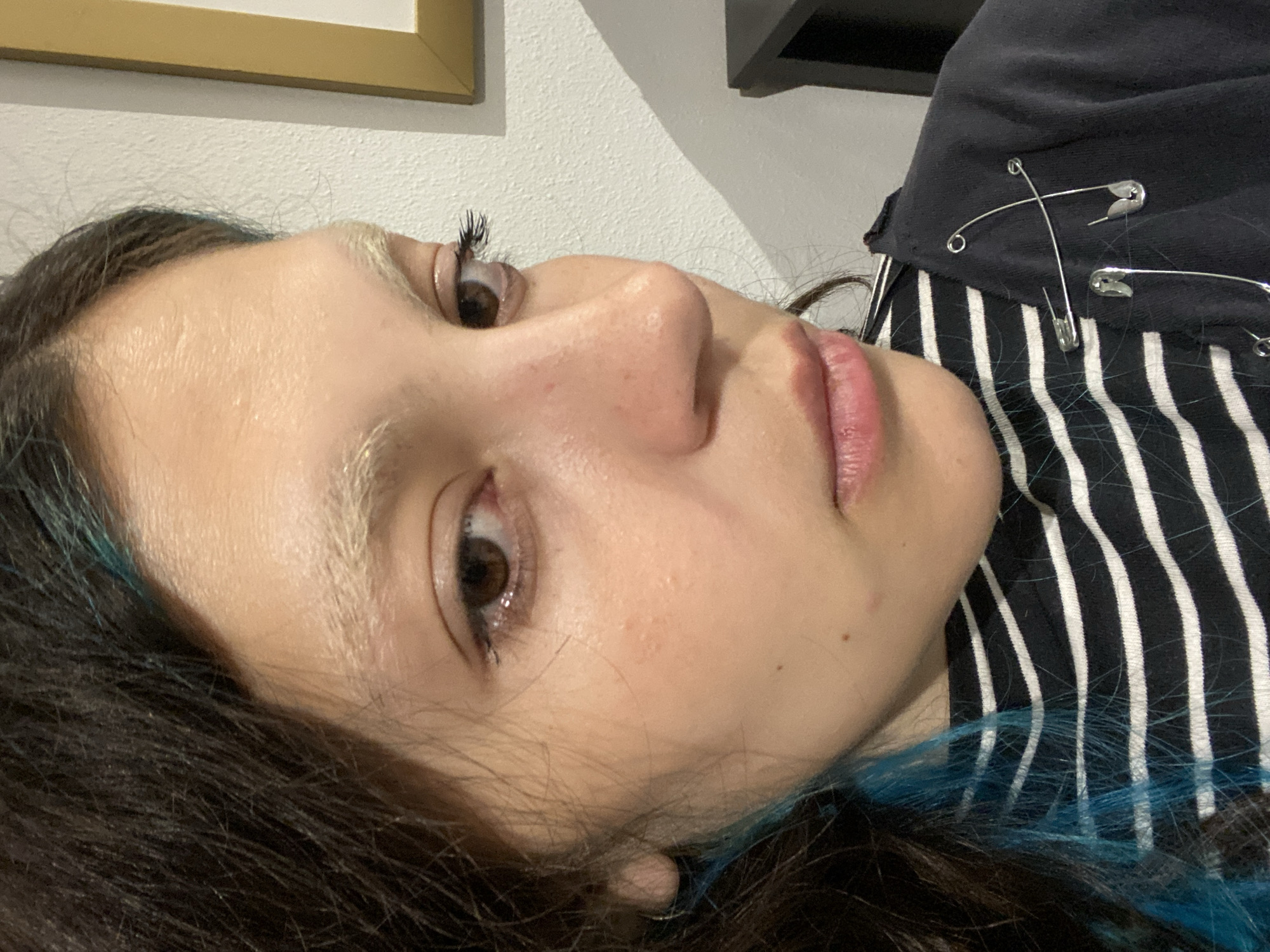A close-up, angled shot of a person's face and upper body. The person has fair skin, light blonde eyebrows, and brown eyes. Their dark brown hair has strands of blue. They are wearing a black and white striped shirt with a black hooded jacket adorned with three safety pins. The background is a light grey wall, with a gold-framed picture visible in the upper left and a dark shelf in the upper right. The location in the city of Gijón/Xixón, Spain is unknown.
