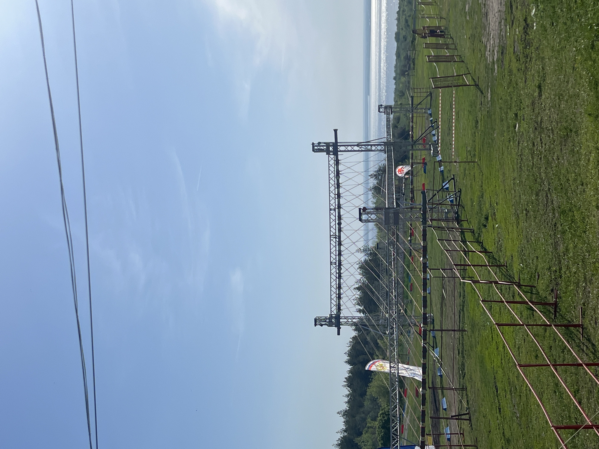 The image depicts an outdoor obstacle course. The course is constructed of metal scaffolding and ropes. The surrounding environment is a grassy field with a treeline in the background. In the distance, a body of water is visible. The location is likely Khvalynsk, Russia. Power lines can be seen on the left. There are red fence posts and white and blue flags lining the course.