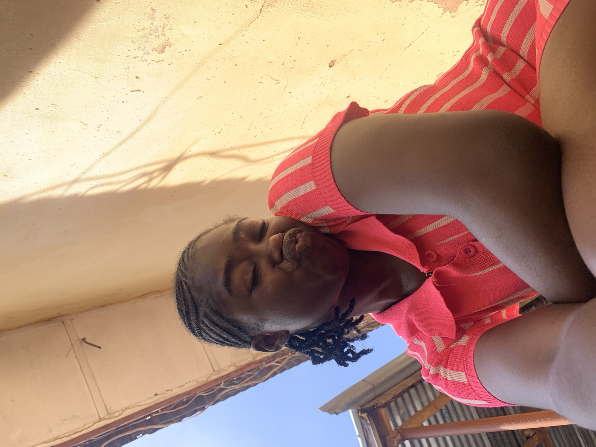 A young individual, appearing female, is depicted in a low-angle, close-up portrait outdoors in Rafin Par, Nigeria. The subject has dark skin and black hair styled in numerous small, tight braids, some hanging loosely near the right ear. The facial expression shows closed eyes and lips intentionally puckered into a prominent kissing shape. The subject wears a short-sleeved, collared knit shirt featuring alternating horizontal stripes of bright coral pink and light beige. Two circular red buttons are visible at the shirt's collar. The subject's right forearm and upper arm, bent at the elbow, are partially visible in the lower foreground. The background consists of a textured, light yellow-ochre wall exhibiting surface irregularities and minor areas of paint peeling. Sharp, elongated shadows, consistent with foliage or thin wires, are cast across the wall from the upper left, indicating strong, direct sunlight. The upper left portion of the image reveals a clear blue sky alongside the edge of a corrugated metal roofing structure, supported by visible wooden beams, with some exposed wiring or metallic rods underneath.
