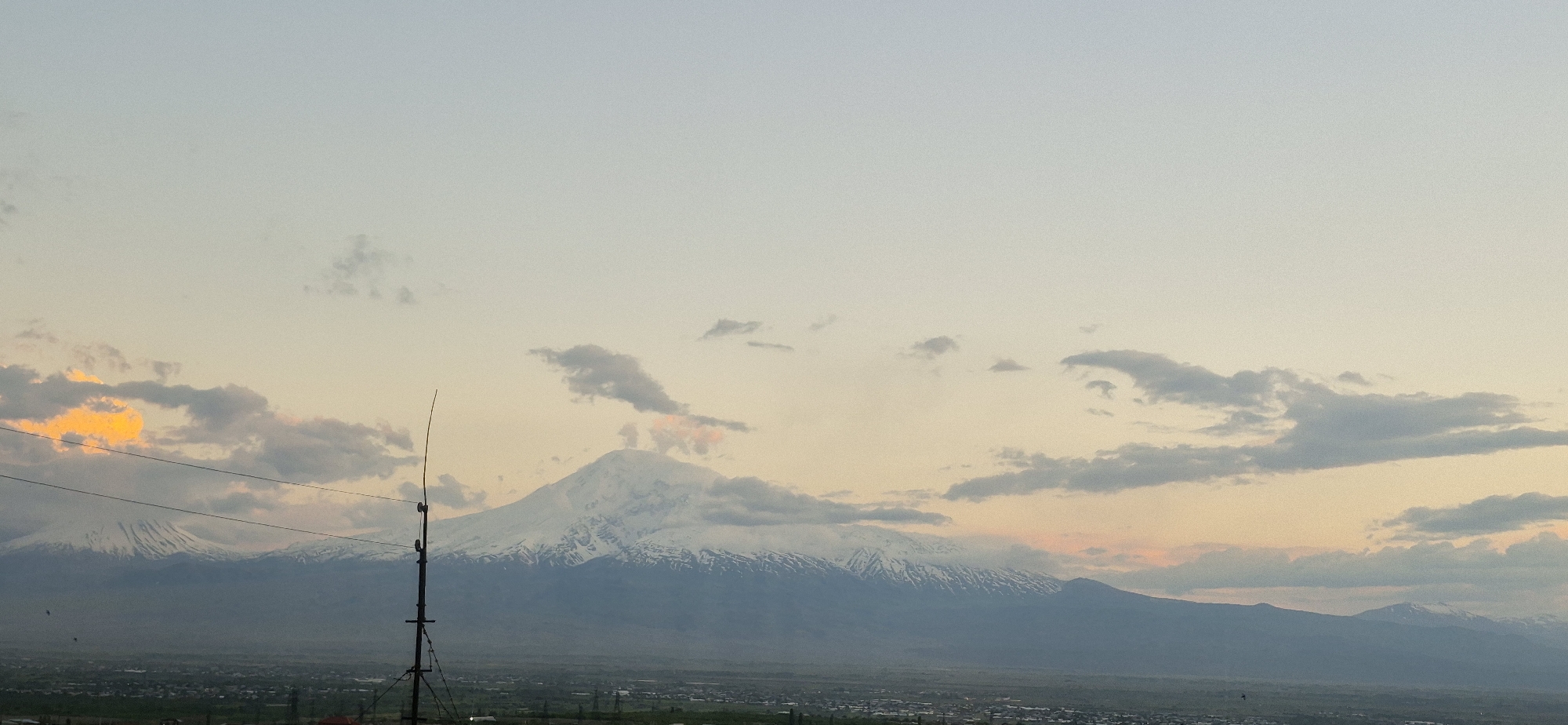 The image shows a landscape with a snow-capped mountain range in the background, possibly at dusk or dawn, given the soft light and cloud formations. A city or town lies in the foreground, stretching into the distance. An electrical pole or antenna stands prominently in the middle ground, with wires visible near the top left corner. The location is Unknown, Unknown.