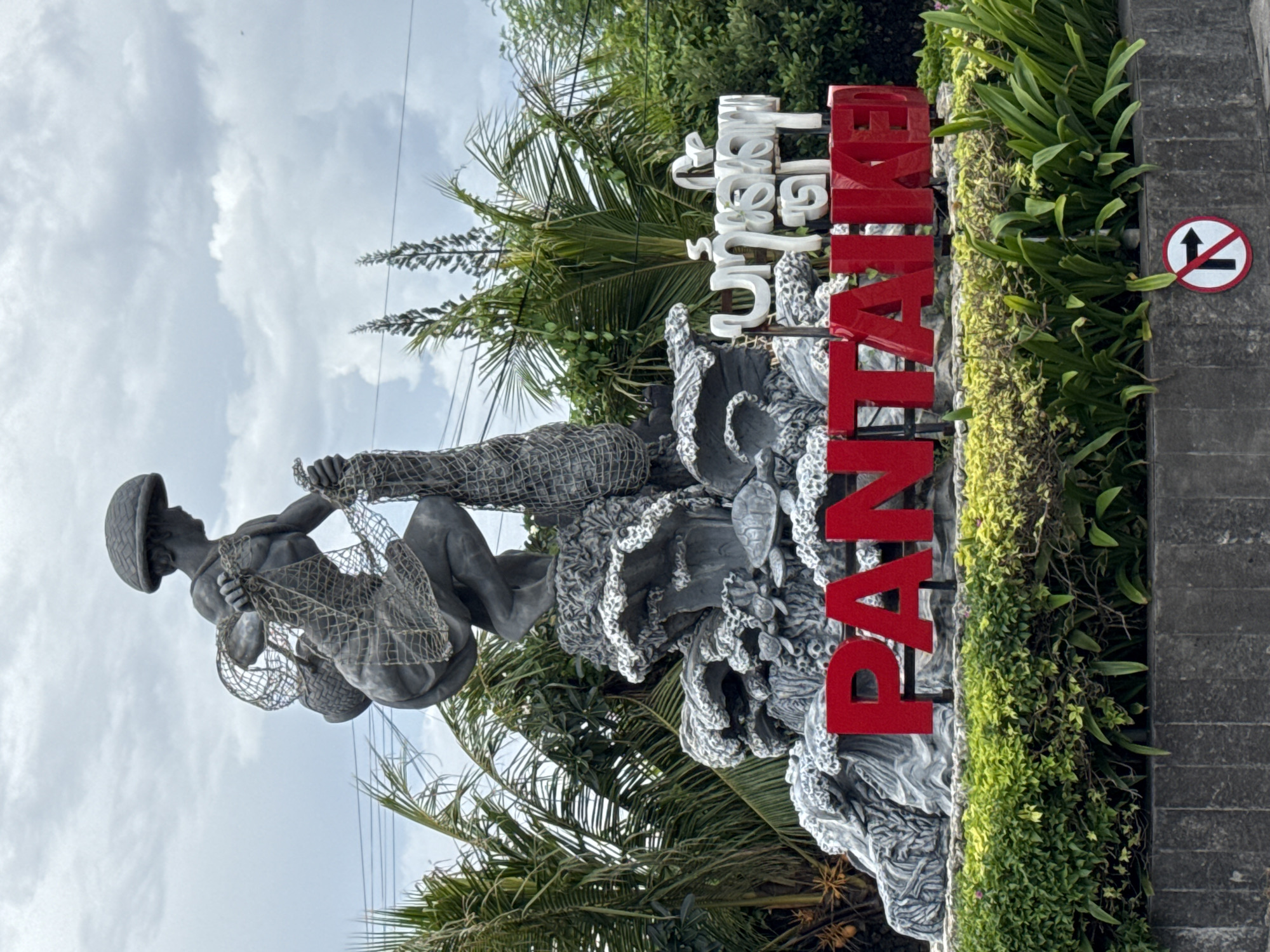 The image depicts a statue of a fisherman set against a backdrop of palm trees and a partly cloudy sky. The statue, likely made of stone or a similar material, shows a man in traditional attire and a hat, holding fishing nets. Adjacent to the statue, a sign reads "PANTAI" in large red letters, with smaller, white text above in a local language. The arrangement is set within a landscaped area, with plants and vegetation framing the base. A traffic sign with a prohibition symbol is visible. The location is in Kuta, Indonesia, indicated by the sign that reads "PANTAI" which translates to "beach".