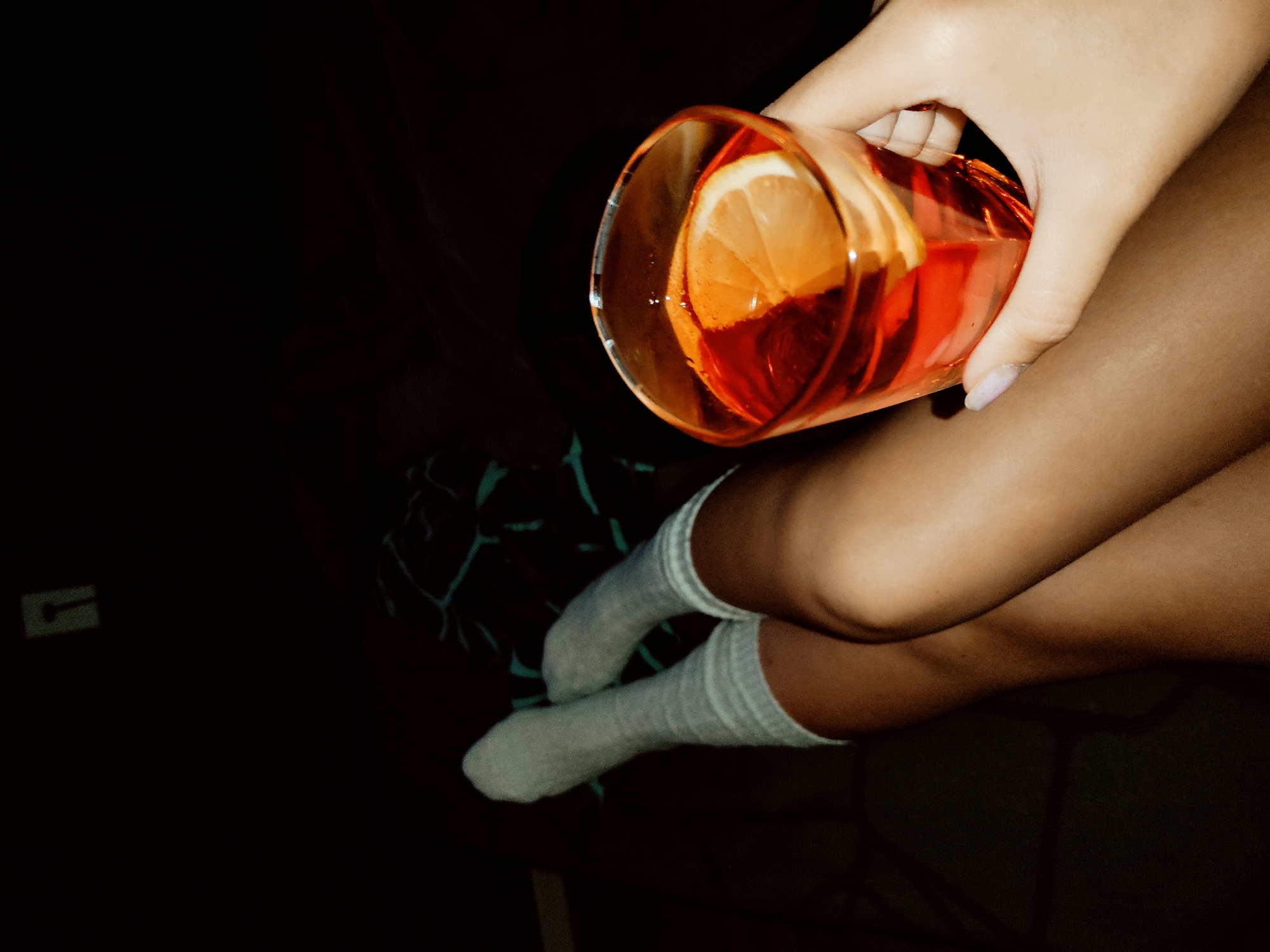 A person is holding a glass of amber-colored liquid with a lemon slice while sitting with crossed legs. The person is wearing white socks. The background is dark, obscuring details, but a bed or chair with a dark patterned fabric is partially visible. There is a dark rectangle in the lower left corner.
The city of Chaikovsky, Russia, is not discernible in the image.