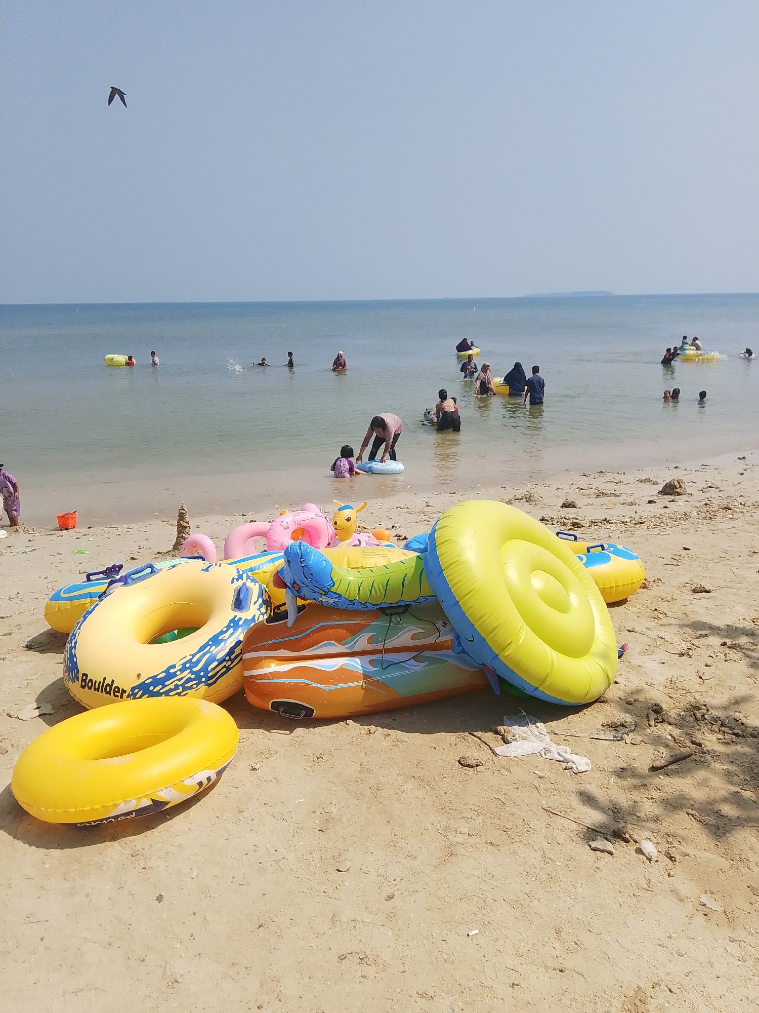 The image depicts a beach scene in Pejagan, Indonesia, with the calm sea in the background and a sandy shore in the foreground. The sky is clear and pale blue. Several inflatable tubes and rafts are scattered on the sand, with a larger, yellow and blue striped tube prominently placed in the lower left corner. In the water, numerous people, including adults and children, are visible. Some are swimming, while others are using inflatable devices. A bird is seen flying in the upper left quadrant of the image. The overall atmosphere suggests a recreational outing at the beach.