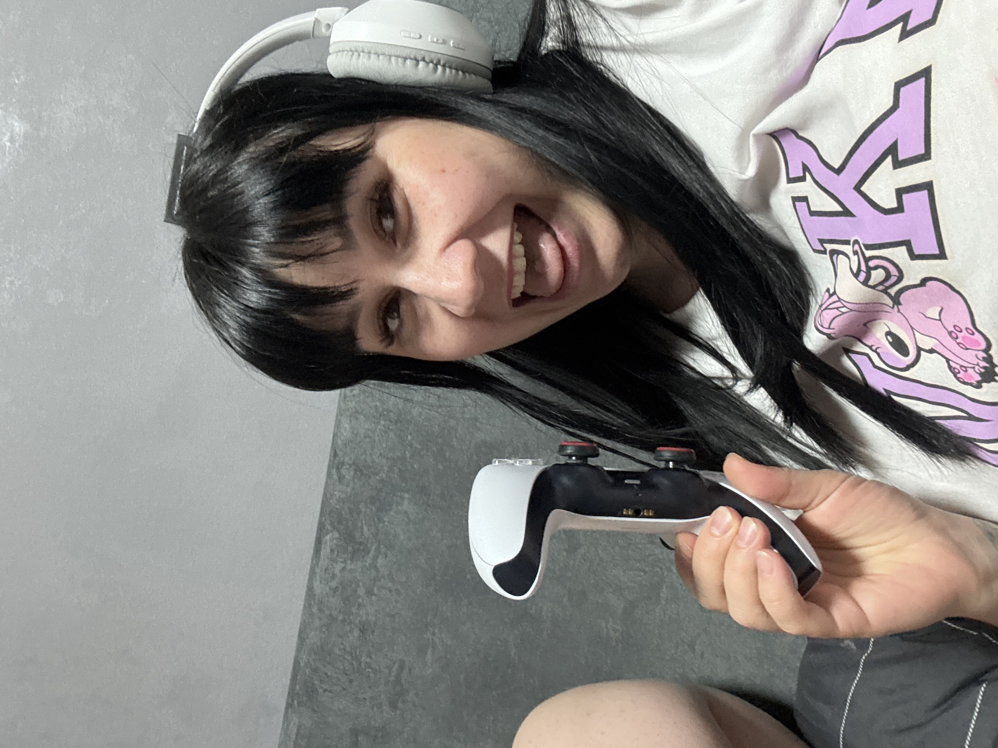A young woman is holding a PlayStation 5 controller in her right hand. She is smiling with an open mouth, revealing her teeth. She has black hair with bangs and white headphones on her head. She's wearing a white t-shirt with a purple design. The background appears to be a gray wall. No information related to Zav'yalovo, Russia is present.
