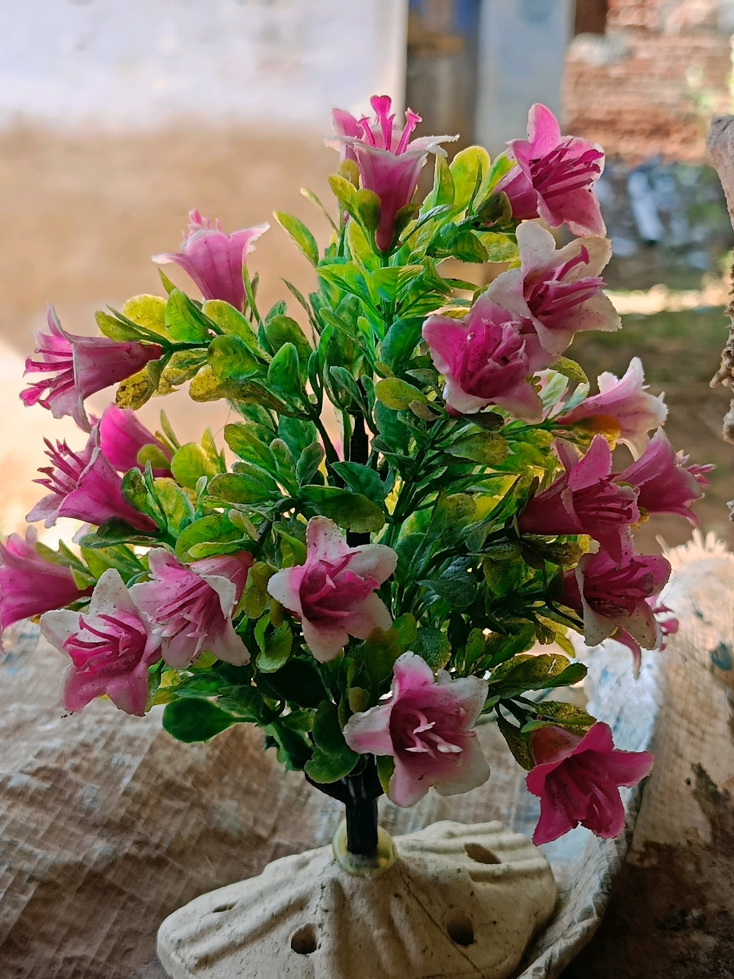 This is a close-up, eye-level, outdoor, artificial light photograph of a bouquet of pink and white artificial flowers with green leaves. The flowers have trumpet-shaped blooms with prominent stamens. They are arranged in a bunch and supported by a dark stem emerging from a beige, roughly textured decorative vase that appears to be made of concrete or ceramic. The vase has a few small circular holes.
The flowers and vase are placed on a textured, light brown surface that resembles burlap or a rough cloth. In the background, the scene is out of focus, showing a mottled light brown and beige wall on the left, and a blurred brick wall and structure on the right. There is no visible sign of the city of Tirunelveli, India, in the image. The image depicts a static, still-life arrangement.