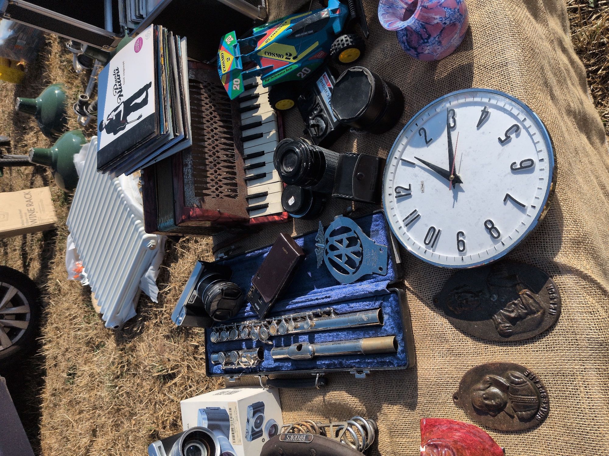 A daylight outdoor market scene, possibly in Claverley, United Kingdom, displays a variety of items laid out on burlap fabric and dry grass. In the background, a car tire and a portion of a vehicle are visible, along with two green industrial-style lamps and a brown cardboard box labeled "WINE RACK."
The foreground is occupied by several distinct objects. To the left, a silver-colored flute is neatly placed inside a dark blue case. Next to it, a white camera box with images of cameras is partially visible. A black camera with a lens is positioned above the flute case. In the center, an accordion with a wooden casing and white keys is set down, with a stack of records, one featuring the artist "Paolo Nutini," resting on top of it. To the right of the accordion, a colorful toy race car with the number "20" and the word "COSMO" printed on it is displayed.
Further down, a large round clock with a white face and black numbers sits prominently. To its left are two oval, dark metal plaques with embossed figures and inscriptions, one reading "SHAKESPEARE." To the far right, a small, decorative glass vase with pink and blue marbling is placed. Additional items include camera lenses and what appears to be a vintage leather bicycle saddle on the left. The ground is covered with dry grass and burlap, suggesting an outdoor flea market or car boot sale. No people are visible in the frame.