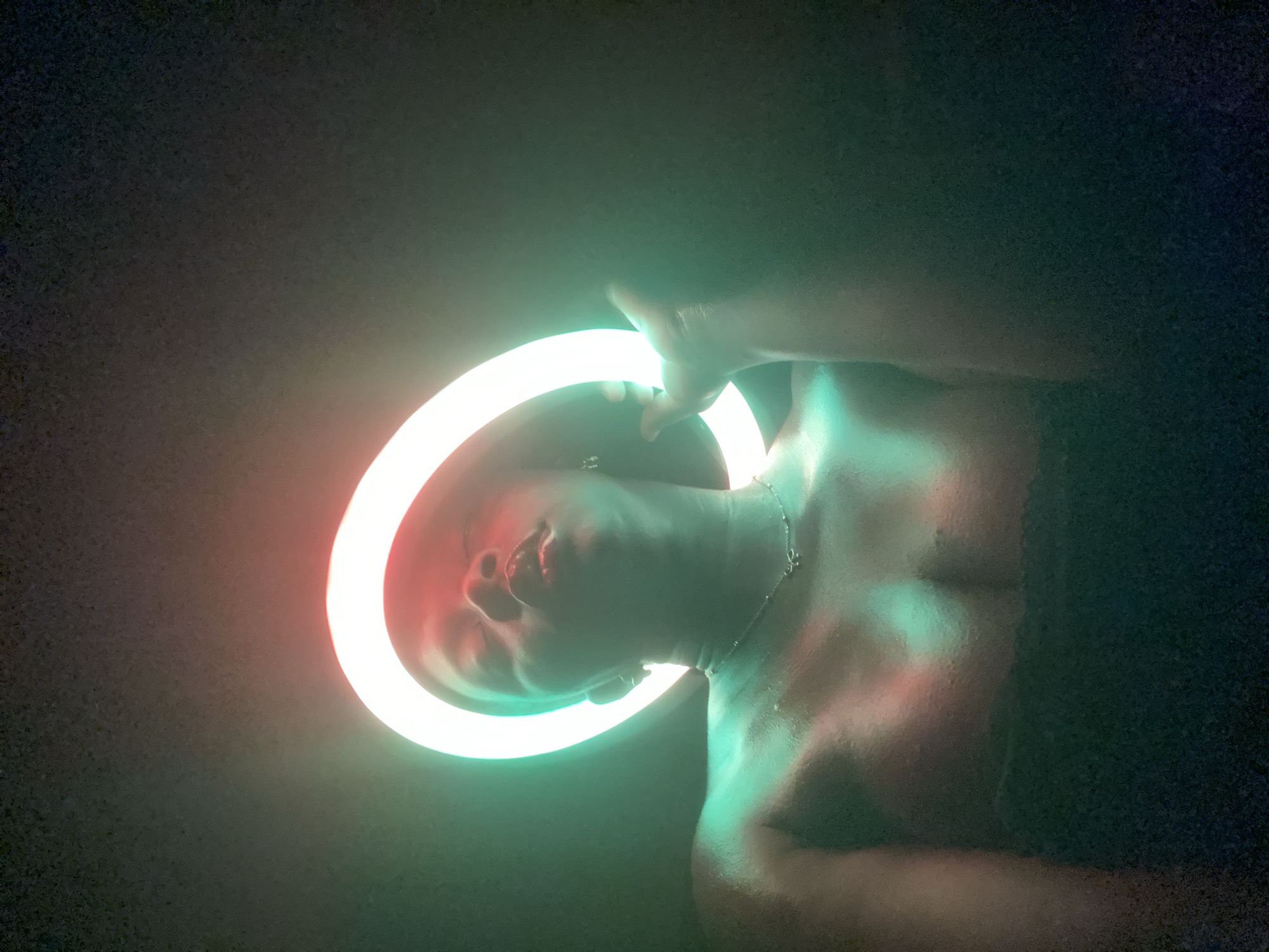 The image features a person, likely an adult, framed from the chest up against a dark, indistinct background. The primary light source is a glowing ring light, which the person holds or interacts with. The light casts prominent red and green hues: green illuminates the person's chest and one side of their face, while red highlights the other side of their face and head.
The person's head is tilted slightly upwards, with their mouth slightly ajar. Their features are partially obscured by the intense, colored light, creating a dramatic and artistic effect. One hand is visible, appearing to grasp or interact with the luminous ring. A delicate silver chain necklace adorns their neck. The person appears to be bare-chested or wearing minimal, dark attire that merges with the shadowy surroundings.
The background is uniformly dark and out of focus, emphasizing the illuminated subject. The overall atmosphere is dark, suggesting an indoor setting or nighttime, with the ring light being the sole or dominant light source. There is no visible text in the image. The scene is set in Maigizo, Nigeria.