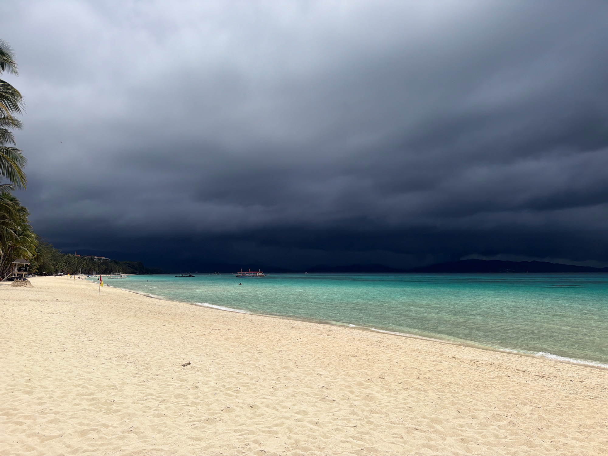 The image depicts a tropical beach scene on a cloudy day, likely located in Boracay, Malay, Philippines. A wide expanse of pale sand stretches from the foreground towards the left, meeting a strip of turquoise ocean. The water is calm with gentle waves lapping the shore. In the distance, several boats are visible on the water, and the shoreline of another island or landmass can be seen on the horizon. The sky is dominated by dark, ominous storm clouds that cast a dramatic shadow over the scene, creating a stark contrast between the dark sky and the bright ocean. A few palm trees are visible on the far left of the image, adding to the tropical ambiance. There are a few people scattered along the beach and in the water, engaged in leisurely activities. The overall impression is one of impending weather change, with the beauty of the beach juxtaposed against the dramatic and foreboding sky.