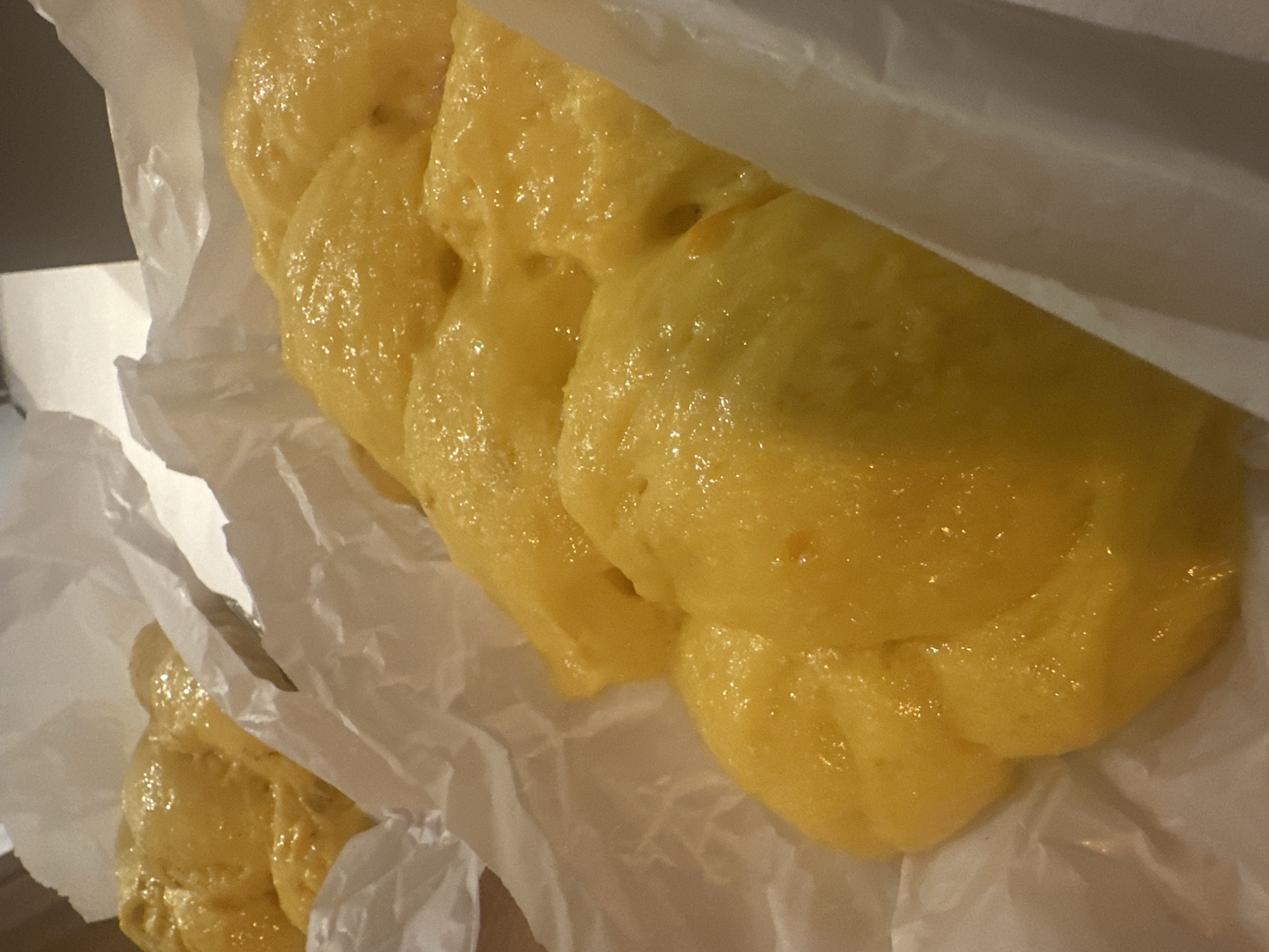 A close-up shot reveals a cluster of golden-yellow, glazed pastries resting on crumpled white parchment paper within a white, possibly plastic, bag. The pastries appear soft and slightly misshapen, with a glossy surface reflecting light. The top right corner of the image is partially obscured by a semi-transparent material, likely part of the bag. The background is blurred and indistinct, but hints at a surface that may be a countertop or table. There is no indication of location or people.
