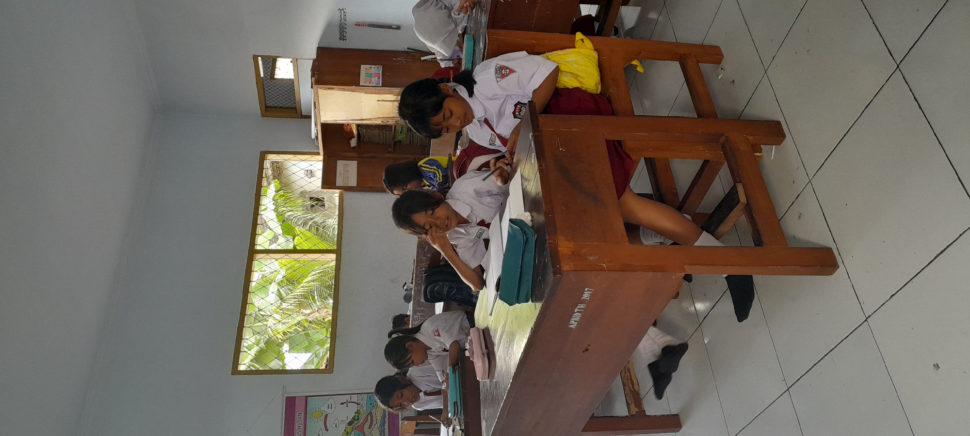 The image depicts a classroom scene in Rogojampi, Indonesia, with several young students seated at wooden desks, engaged in learning activities. The room is well-lit by natural light filtering through a window with a diamond-patterned grate, revealing lush green foliage outside. On the walls, educational posters are visible. The students, predominantly girls, are wearing school uniforms consisting of white shirts and red skirts or shorts. They are seen writing, reading, and interacting with their study materials. Some students have pencils or pens in hand, and books and notebooks are spread across the desks. A wooden cabinet stands against the wall, containing stacked books. The floor is tiled with white squares. The desks themselves are also made of wood, and one desk has "APBD TH. 2017" inscribed on its side. The overall atmosphere suggests a typical school day in a primary or junior high school.