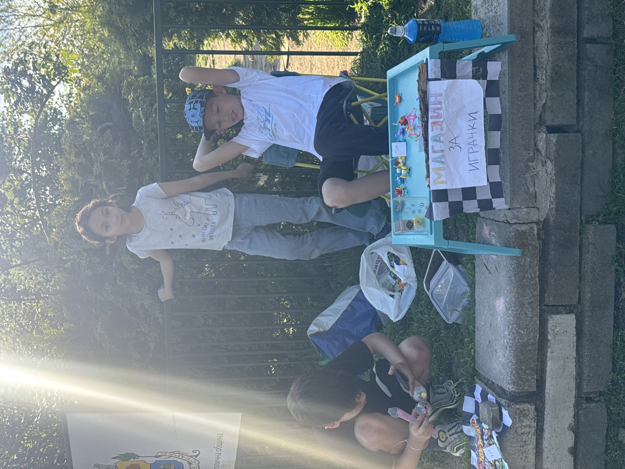 Three children are involved in a toy business. A girl in a unicorn t-shirt leans on a fence. A boy wearing a blue hat and white t-shirt sits on a blue plastic chair behind a blue plastic table. Toys for sale are on the table. A hand-written sign on a checkered piece of fabric reads "МАГАЗИН ЗА ИГРАЧКИ" (Shop for Toys). A plastic bag with more toys and a blue bottle of water are present. A third child is crouched down on the grass near a stone border and is holding a toy. The location is outdoors in Sofia, Bulgaria.