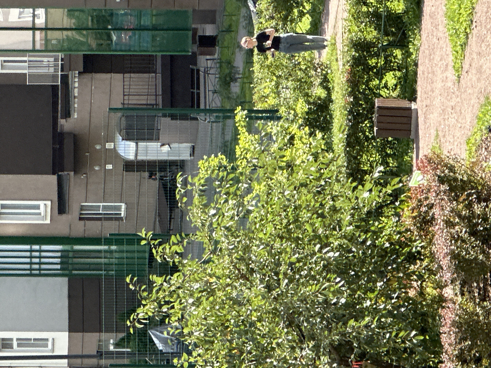 In the city of Unknown, Russia, an outdoor scene captures a woman walking on a path through a landscaped area. She is wearing a black t-shirt and grey pants and is holding a dark-colored cup in her right hand. The environment features lush green trees and bushes, with a fence visible in the background. Beyond the fence, a building with multiple windows and balconies is seen, with green architectural elements and a light-colored facade. A brown trash receptacle stands on the side of the path. The ground is covered with gravel and grass. The lighting suggests it is daytime, with sunlight filtering through the foliage.
