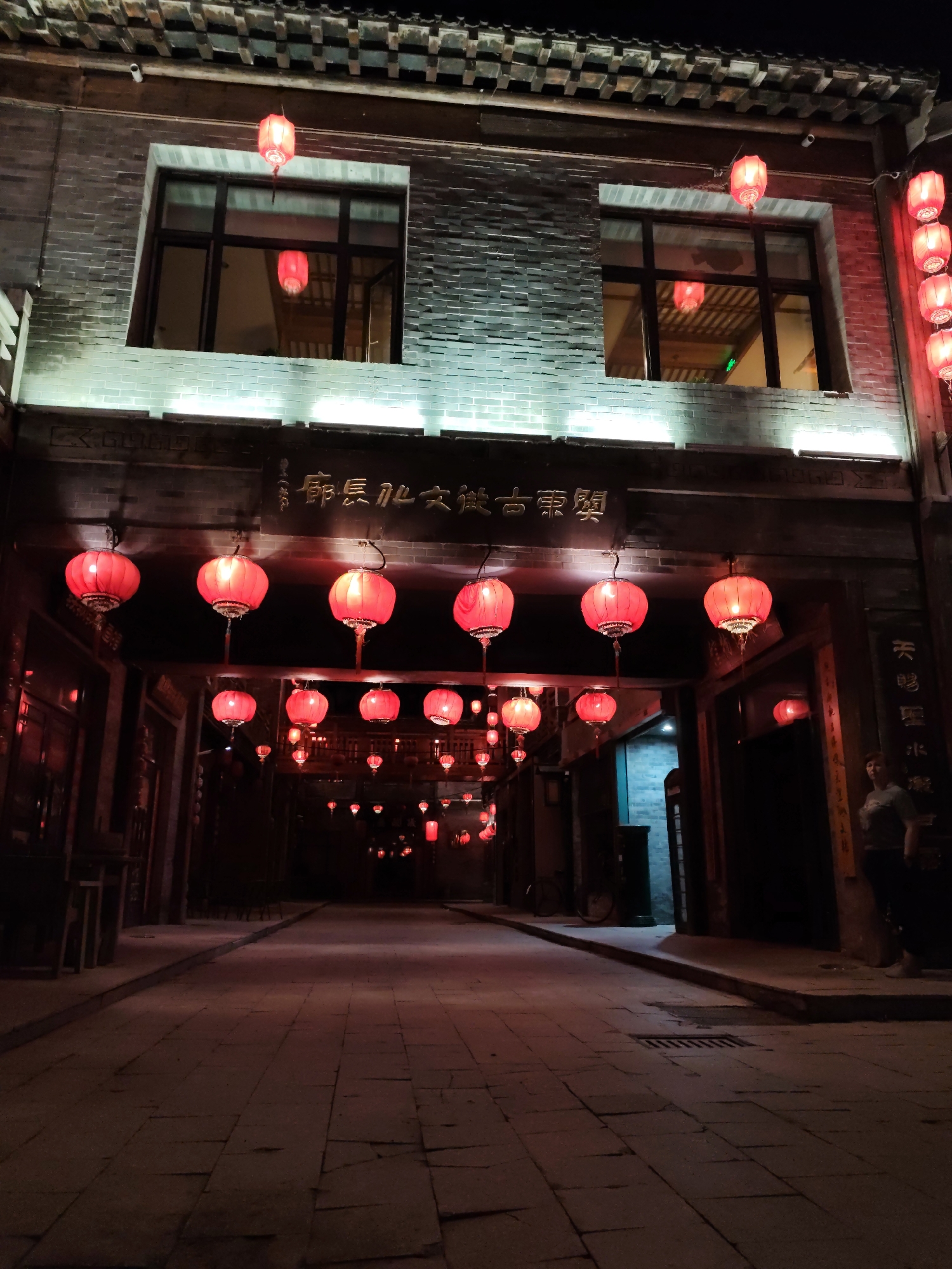 The image depicts a nighttime scene on a cobblestone street in Lindian, China, likely a historic or culturally significant area. Red lanterns are prominently displayed, illuminating the narrow street and the traditional architecture of the buildings on either side. The buildings appear to be made of brick with tiled roofs, and feature large windows. One building has Chinese characters inscribed above an archway. A few people are visible, including one individual standing near a doorway and another in the distance. The overall atmosphere is festive and traditional, suggesting a special event or a lively evening in the city.