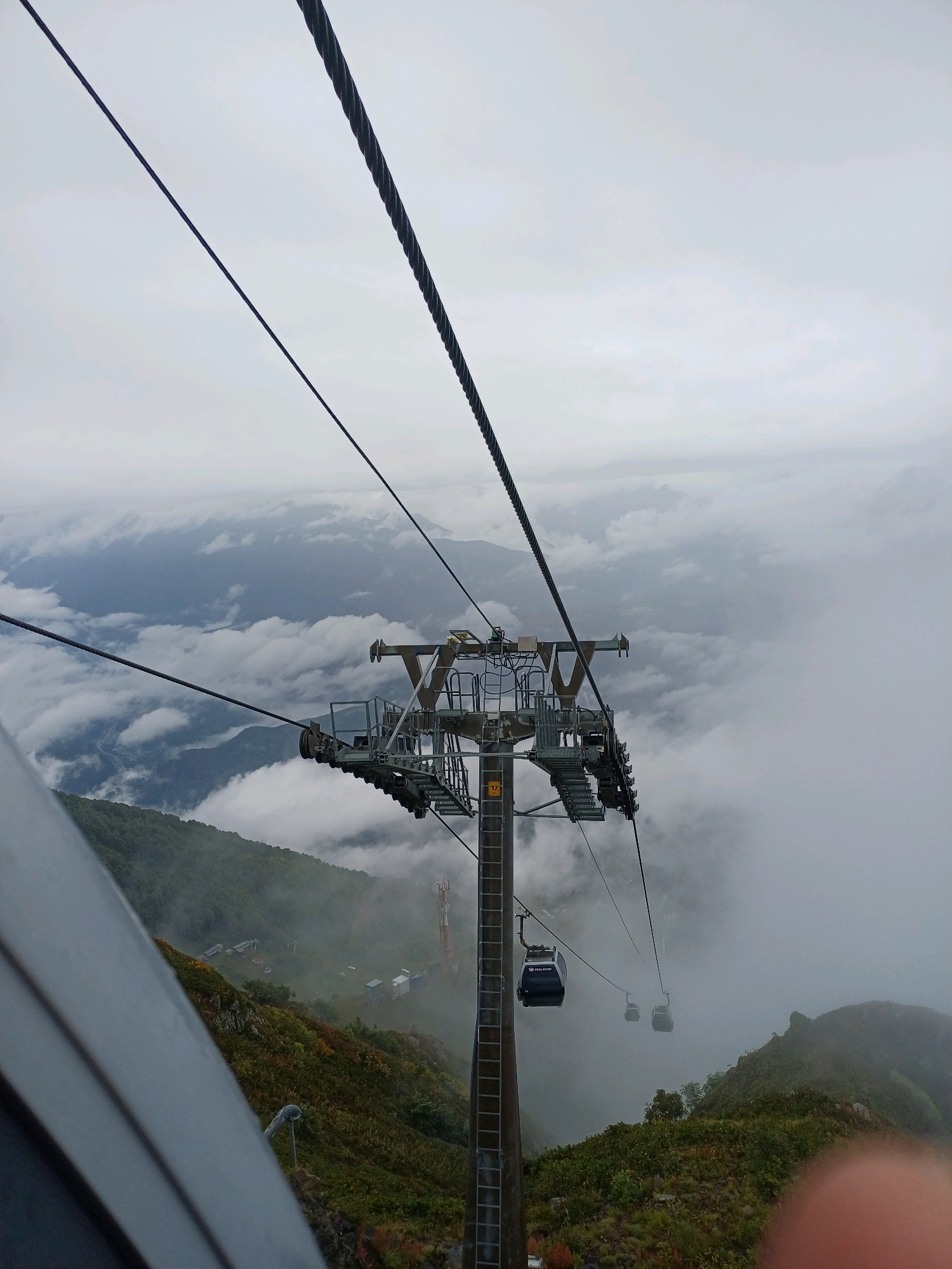The image depicts a scene from a cable car in Estosadok, Russia, looking down at a misty mountain landscape. Several cable cars are visible, suspended by thick steel cables. One cable car is closer to the foreground, displaying the text "ROSA SITOP". Below, the mountainous terrain is covered in green vegetation and shrouded in clouds. A tall communication tower is also visible in the distance. The sky is overcast with heavy clouds, contributing to the overall misty and atmospheric conditions. The perspective suggests the viewer is inside another cable car, looking outwards and downwards.