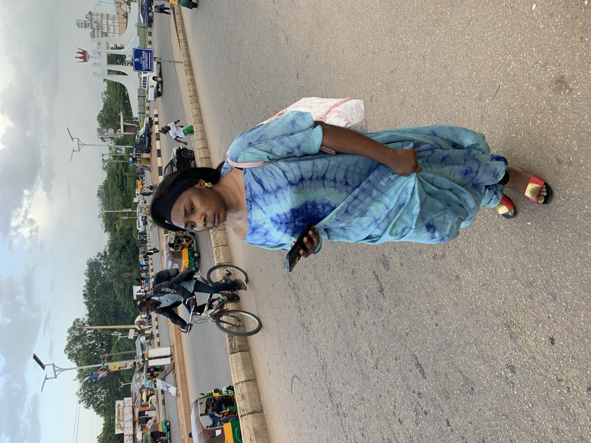 In Jos, Nigeria, a woman in a tie-dye blue dress walks on a street, holding a smartphone. She is wearing red sandals with gold accents. In the background, a man cycles a bicycle, and a tuk-tuk is visible. A distinctive white sculpture with a crown at the top, a landmark in Jos, stands in the distance, along with other buildings and trees. The sky is cloudy.
