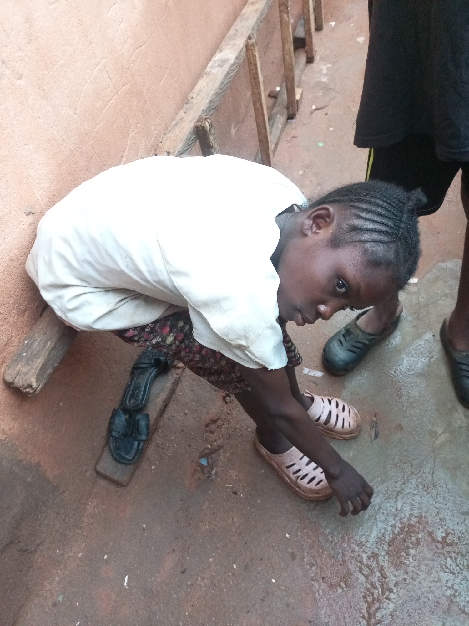 A young girl with braided hair is sitting on a wooden beam that is part of a makeshift structure, possibly a fence or wall, in an outdoor urban environment. She is wearing a white shirt and a patterned skirt, and her feet are clad in light-colored slip-on shoes. A pair of black sandals are placed on a wooden plank near her. The ground is wet and appears to be a dirt or concrete surface, with puddles and debris scattered around. The wall behind her is a textured, peach-colored surface. The overall scene suggests a humble, possibly impoverished, setting in Tempure, Nigeria.