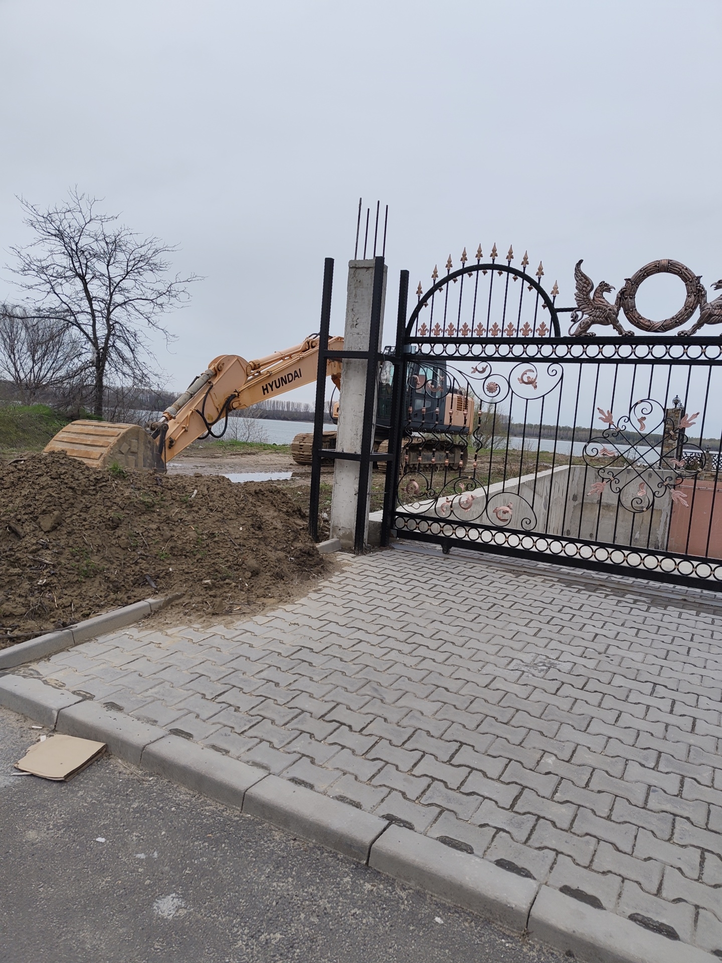 An excavator is positioned near a decorative metal gate in Kozloduy, Bulgaria. The excavator's bucket is next to a pile of dirt. The foreground features gray paving stones and a curb. Behind the gate, a body of water is visible. The sky is overcast.