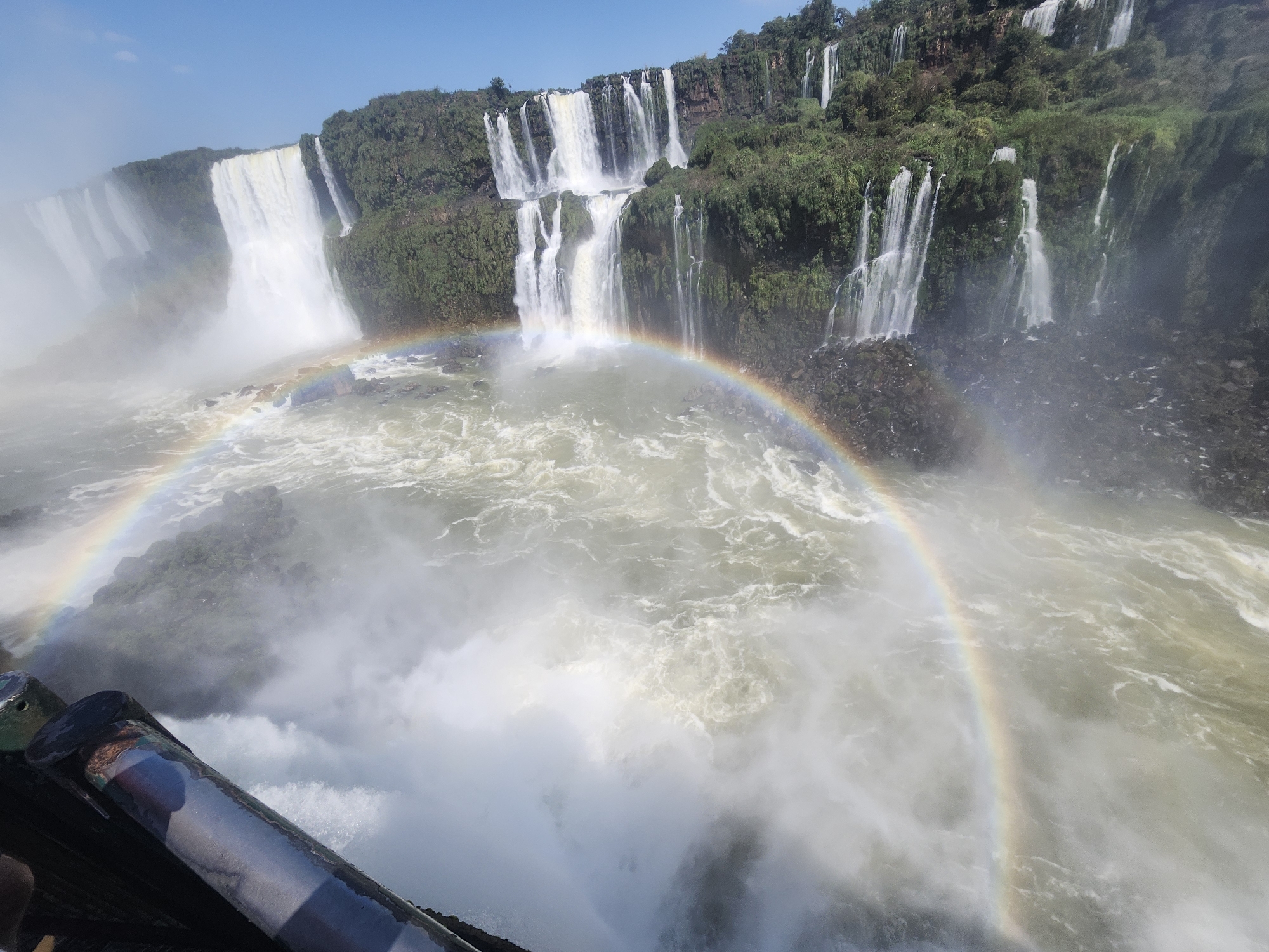 The image shows the Iguazu Falls in Foz do Iguaçu, Brazil, a stunning natural wonder characterized by a series of cascading waterfalls. The water plunges dramatically over a rocky cliff face covered in lush green vegetation. A vibrant rainbow arches across the mist-filled air above the churning water at the base of the falls, indicating sunlight interacting with the spray. In the foreground, a section of a railing or barrier, possibly part of a viewing platform, is visible, suggesting a perspective from a tourist area. The sky above is clear and blue.