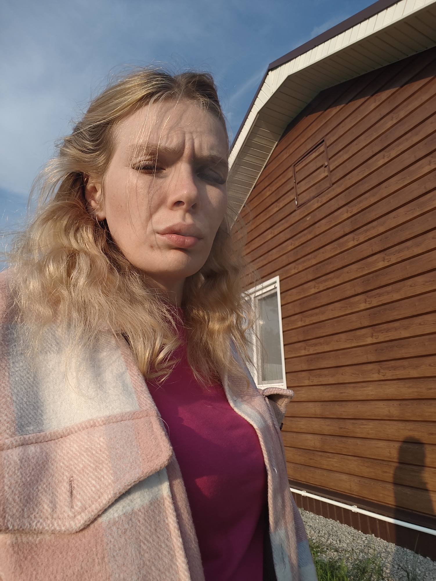 A blonde woman with wavy hair is captured in a selfie taken outdoors in front of a wooden building. She is wearing a pink t-shirt and a pink and white plaid jacket. Her face is partially obscured by her hair, and she has a slightly pensive expression. The background shows the side of a brown wooden structure with a white-trimmed roof and a window. The sky above is blue with some clouds. The ground in the foreground appears to be covered with gravel and some green grass. The image suggests a sunny day, likely in a rural or suburban setting.