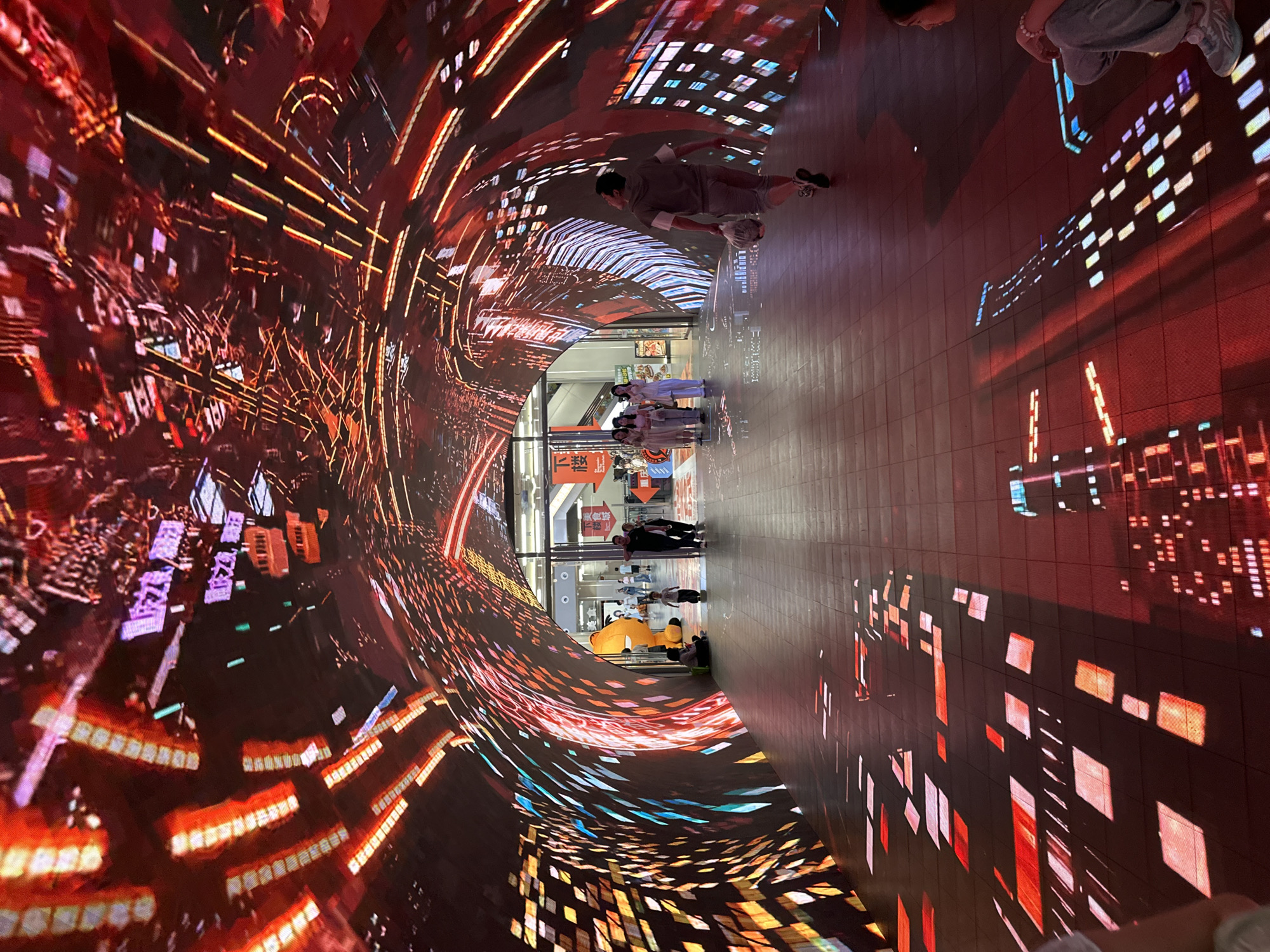 This is a photograph taken indoors, likely in a tunnel or passageway within the city of Yuzhong District, China. The interior of the tunnel is lined with digital screens displaying an animated cityscape with streaks of light that convey a sense of speed or motion. The tunnel floor is tiled, reflecting the light and images from the screens, enhancing the immersive effect.
Several individuals are seen within the tunnel:
- One person is lying on the floor, seemingly posing for a photo or interacting with the visual display.
- A group of people in uniform, possibly staff members, are standing near the exit.
- Other individuals are walking through the tunnel or standing to the side, observing the scene.
The overall environment suggests a modern, urban experience, with the use of digital technology to create an interactive and visually engaging space.