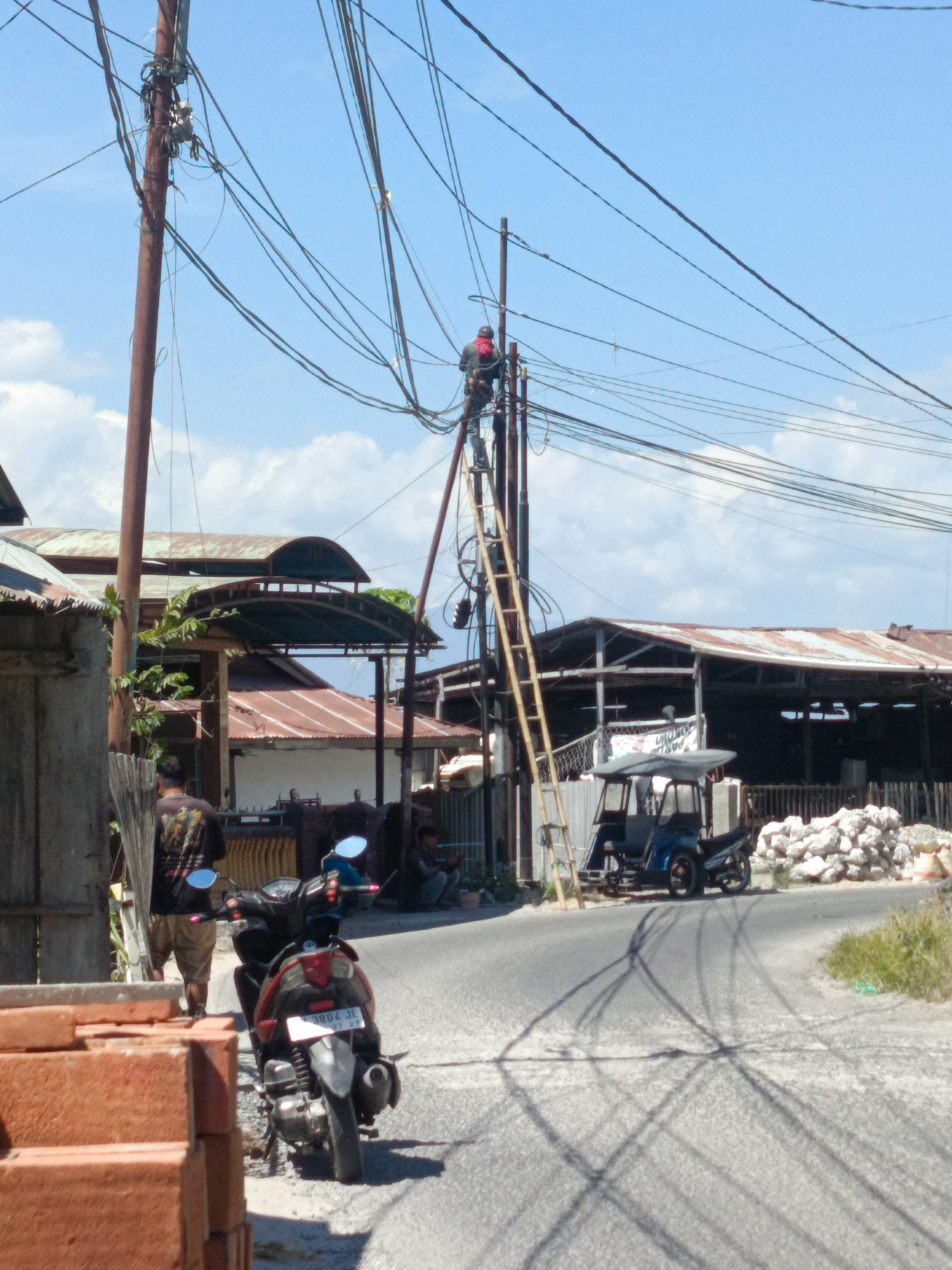The image depicts an outdoor scene in Gorontalo, Indonesia, characterized by a network of overhead power lines. A person, wearing a red shirt and black pants, is positioned on a wooden ladder, actively working on the electrical infrastructure. Below the power lines, a street is visible with a parked motorcycle and a three-wheeled vehicle. The background includes buildings with corrugated metal roofs and a pile of rocks. The overall environment suggests a somewhat utilitarian or industrial area within a town or city.