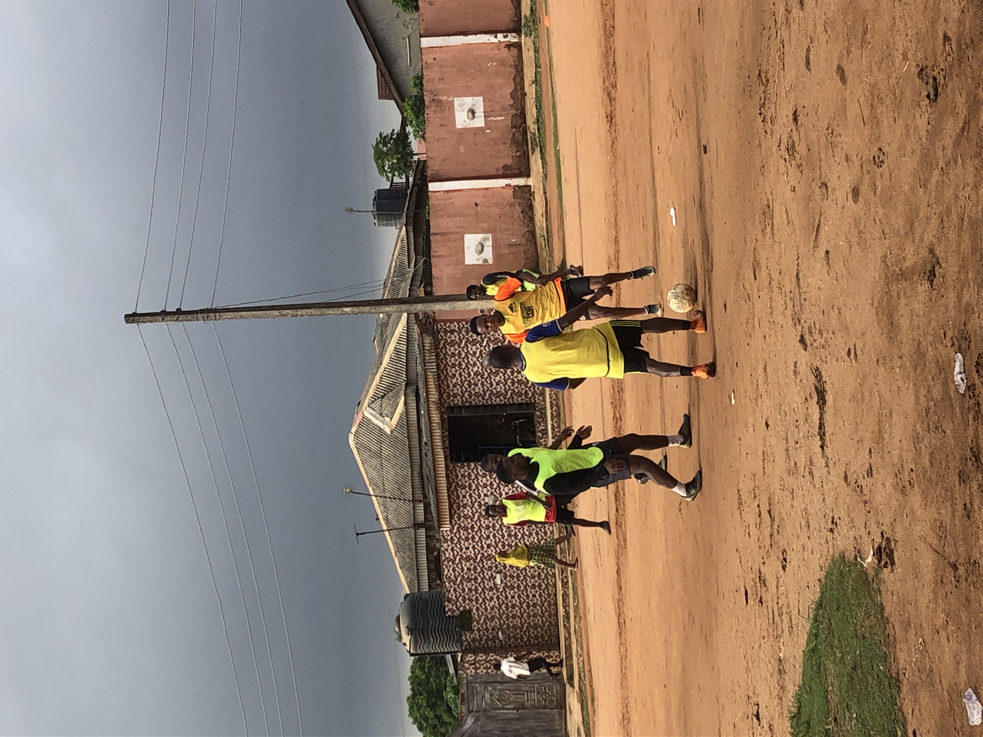 Four people are playing soccer on a dirt field. Two of the people are wearing yellow vests. One person is wearing a green vest. The field is bordered by buildings and an electrical pole. The sky is overcast.