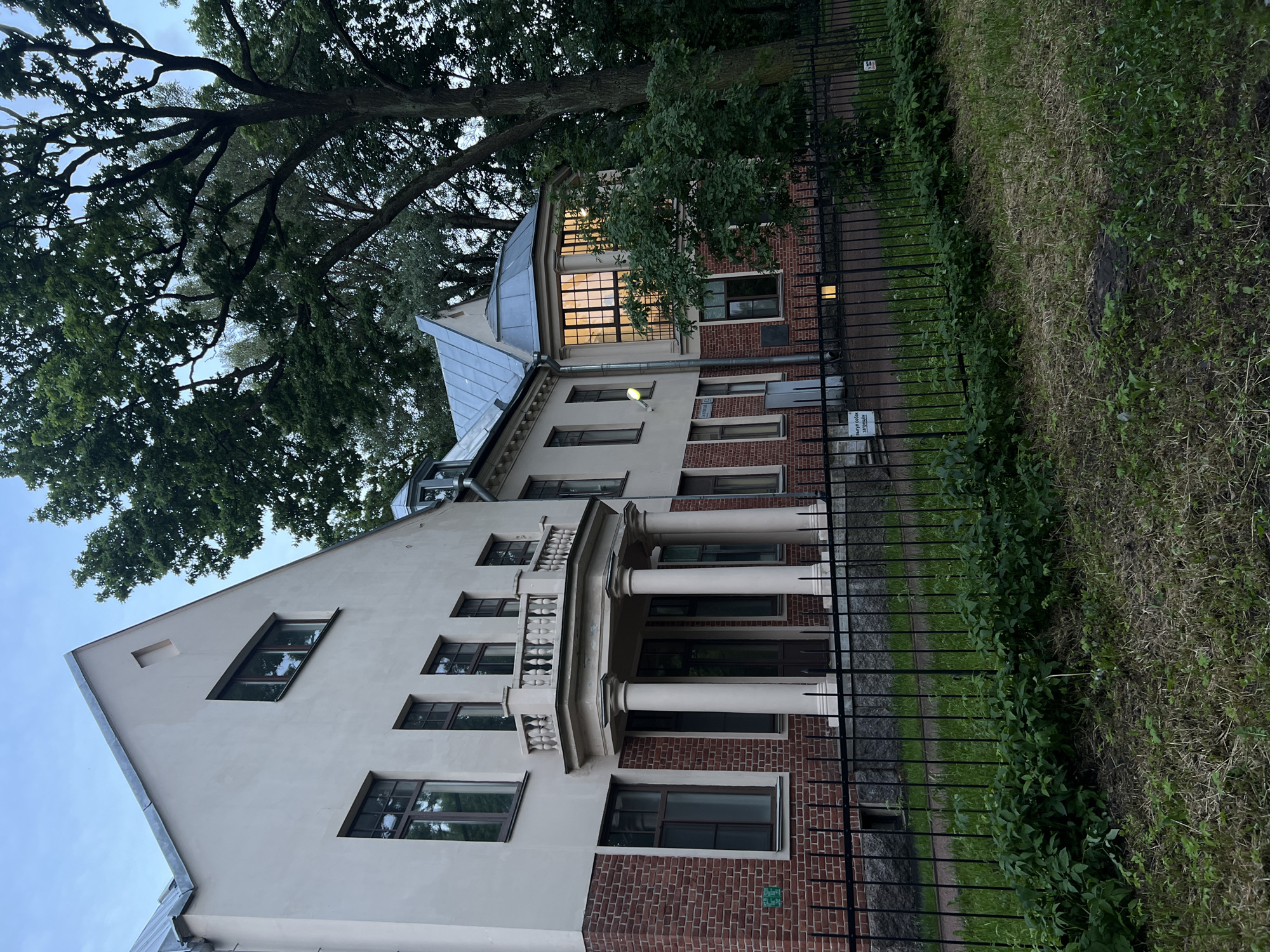 The image depicts a historic building in Saint Petersburg, Russia, specifically the former apartment of architect Alexander Bruni, located at Liteynyy Prospekt, 11. The building, painted a pale yellow with red brick accents on the lower floors, features a prominent portico with four classical columns supporting a balcony. A black wrought-iron fence separates the building from a grassy area and a sidewalk. Mature trees surround the property, casting shadows on the facade and the surrounding grounds. The sky is overcast, suggesting either early morning or late evening. The scene is devoid of any people, and the overall atmosphere is one of quiet stillness, characteristic of a historic architectural site.