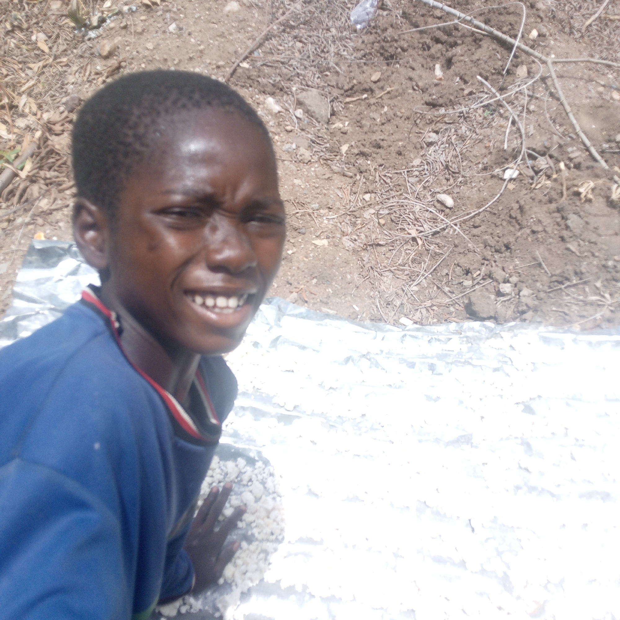 A young Black African boy kneels on a reflective surface. The surface is covered with a layer of what appears to be dried cassava flakes. The boy smiles, and his left hand rests on the layer of flakes. Behind the boy and the reflective surface is a patch of dry, bare ground with scattered twigs and rocks. The location is possibly Adankolo, Nigeria, based on provided information.