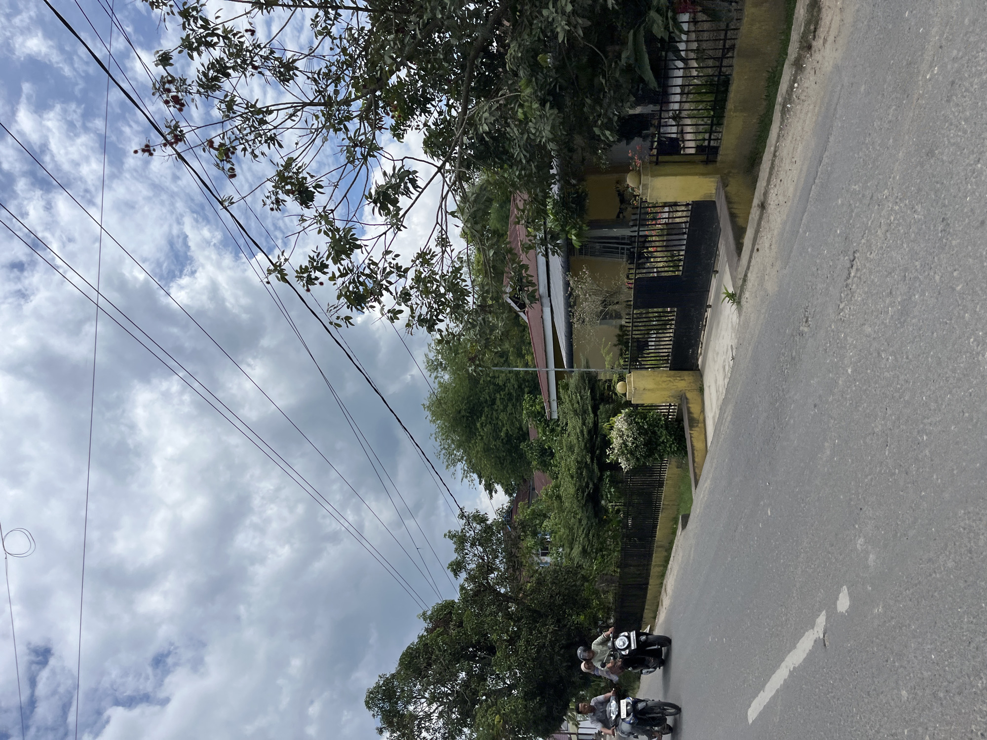 In Langsa, Indonesia, this image captures a street scene with overcast skies. A paved road with a white dashed line runs through the foreground. Two motorcycles are visible on the road, with riders. The motorcycle on the left carries two individuals, while the motorcycle on the right carries a single rider.
To the right of the road, a property with a yellow facade and a red-tiled roof is partially obscured by dense foliage. The property is bordered by a black metal fence with yellow posts. A tree with reddish fruit, possibly rambutan, stands near the fence. Power lines crisscross the sky above the scene. The overall impression is a quiet, residential street in a tropical climate.