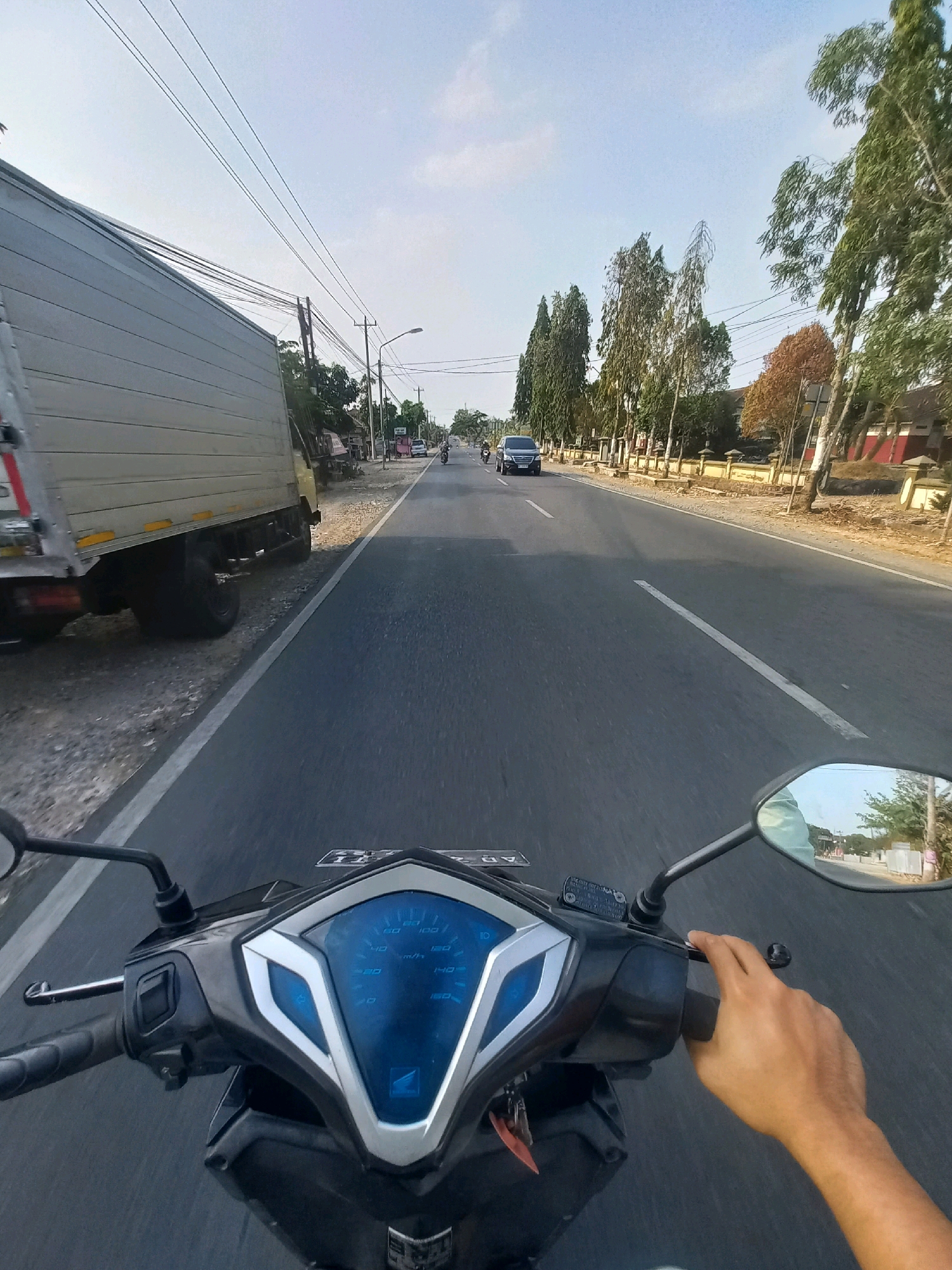 The image depicts a first-person perspective of someone riding a motorcycle on a paved road in Baturetno, Indonesia. To the left of the road, a large, grey box truck is parked or moving slowly. The road itself is asphalt with a white line marking the lane. On the right side of the road, there are trees, some with sparse foliage, and a wall or fence along a property. Power lines stretch across the sky. In the distance, other vehicles, including a dark-colored SUV and several motorcycles, are visible on the road. The sky is a clear, pale blue. The motorcycle's dashboard, with its blue digital display and silver accents, is prominently in the foreground, and a human hand is visible on the right handlebar, appearing to be operating the throttle. The scene suggests a typical day on a road in a semi-urban or rural Indonesian setting.
