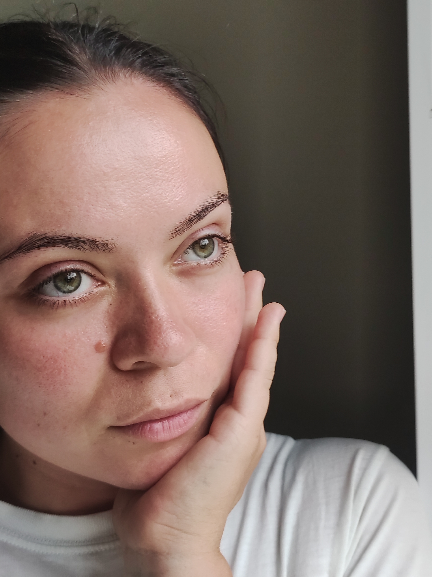 The image is a close-up, slightly angled portrait of a young woman's face and upper chest. She has fair skin with visible pores and some redness on her cheeks. Her eyebrows are dark and well-defined, framing her green eyes which are looking slightly off-camera. She has dark hair pulled back. A small, light brown mole is visible on her left cheekbone. Her right hand is resting on her chin and cheek. She is wearing a white t-shirt. The background is dark and appears to be an indoor setting, possibly near a window, with a sliver of white on the far right edge of the frame. The image does not contain any information about the location in the city of Фоки, Russia.