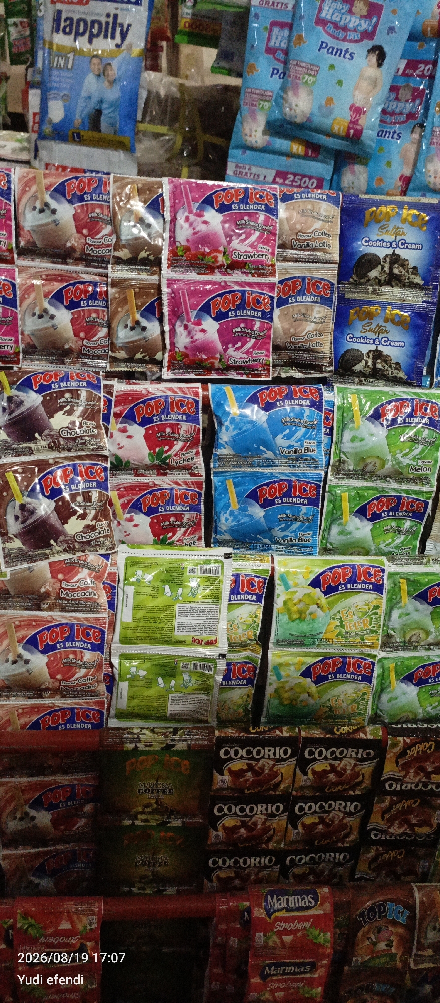 The image displays a retail display of various packaged food and beverage items, predominantly in sachets. The display is densely packed, with multiple rows and columns of products. Dominant brands visible include "Pop Ice" (in various flavors like Strawberry, Cookies & Cream, Vanilla Blue, Lychee, and Melon), "Happily" (possibly a milk powder), "Cocorio," and "Marimas" (Strawberry flavor). There are also some less discernible products in the background. The products are arranged on shelves or a similar display structure. The lighting suggests an indoor environment, likely a small shop or a market stall. The overall impression is one of a well-stocked convenience store or warung. The bottom left corner of the image contains a timestamp "2026/08/19 17:07" and the text "Yudi efendi." This suggests the image was taken in Rawajitu Selatan, Indonesia, possibly at a shop operated or visited by Yudi Efendi, on August 19, 2026, at 5:07 PM.