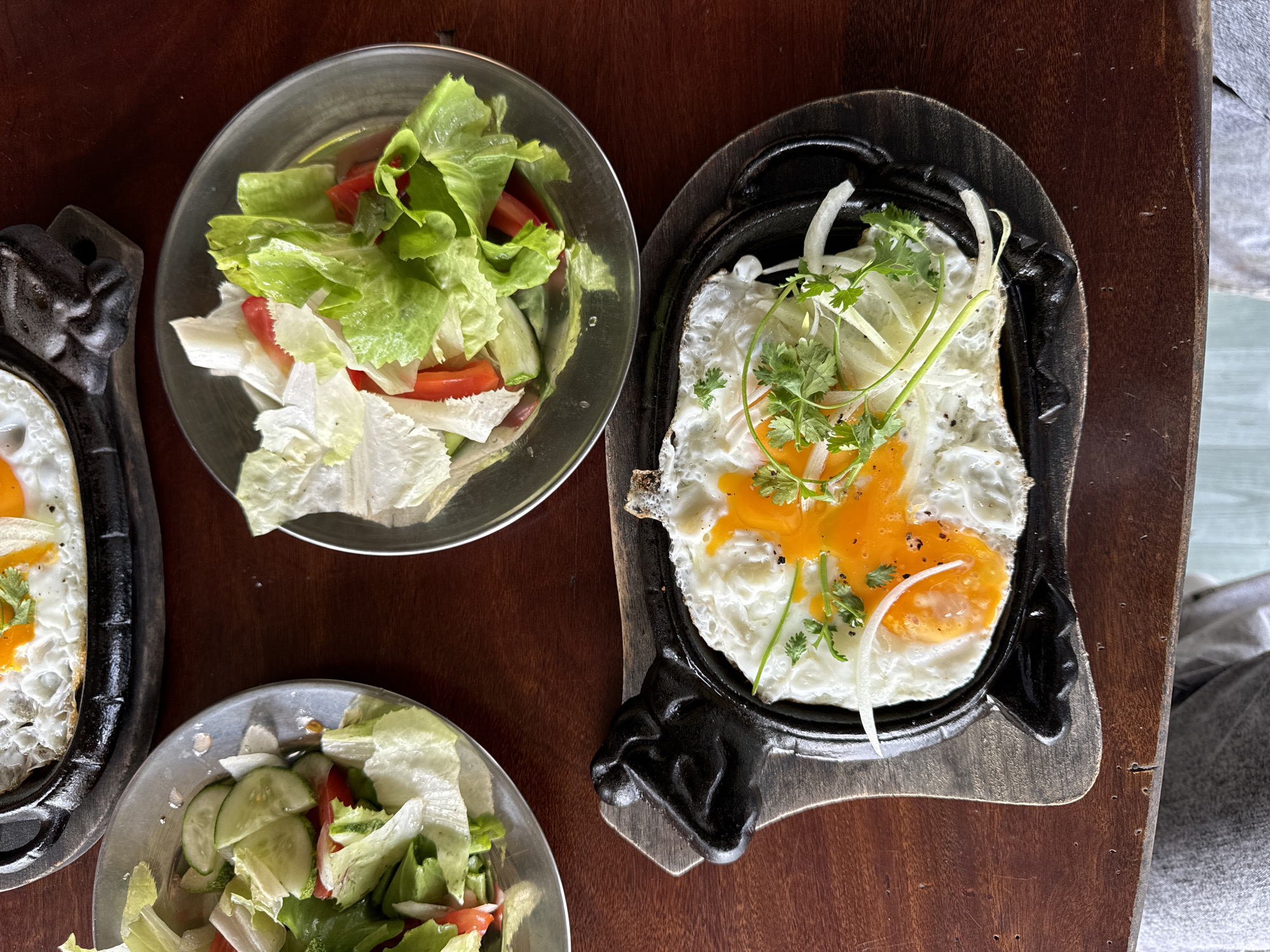 The image presents an overhead view of a meal served on a dark brown wooden table, likely in Sơn Điền, Vietnam. The scene features two individual servings, each including fried eggs on a sizzling plate and a fresh side salad.
Two servings of sunny-side-up fried eggs are prominently displayed on black, oval-shaped cast iron plates, which are themselves set on wooden bases. The egg yolks are a bright orange, surrounded by cooked white. Both egg dishes are garnished with thinly sliced white onions and fresh green cilantro, indicating a fresh seasoning.
Accompanying the main dishes are two small, round, silver-colored metal bowls filled with salad. Both salads consist primarily of fresh green lettuce and sliced red tomatoes. One of the salad bowls also clearly contains slices of cucumber.
The table itself is made of dark brown wood with a visible grain. In the background, beyond the table's edge to the right, a soft, light gray surface, possibly fabric, is visible, along with a hint of a lighter green or blue area, suggesting an outdoor or relaxed indoor setting. The bright, natural lighting indicates it is daytime, likely morning or early afternoon, suitable for a breakfast or brunch meal. No individuals are visible, but the setup implies a meal prepared for at least two people. There is no visible text in the image.