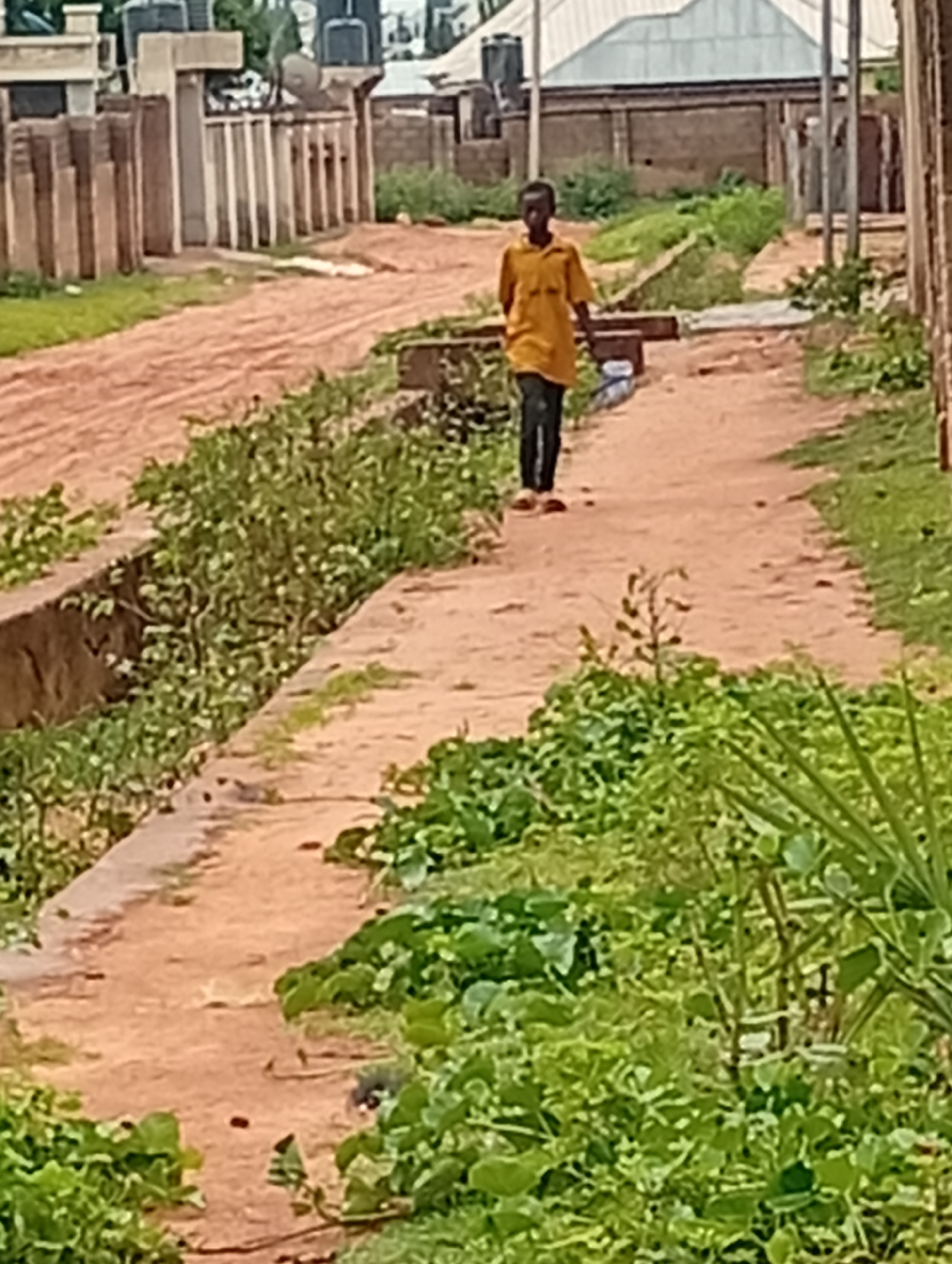 A young boy, appearing to be of African descent, walks on a dirt road in a residential area. He is wearing a yellow short-sleeved collared shirt and black pants. The road is flanked by a ditch on the left and a grassy area on the right. In the background, there are buildings with corrugated metal roofs and a tall fence. The environment is sunny and appears to be outdoors in a village or town. The specific location is Tumfure, Nigeria.