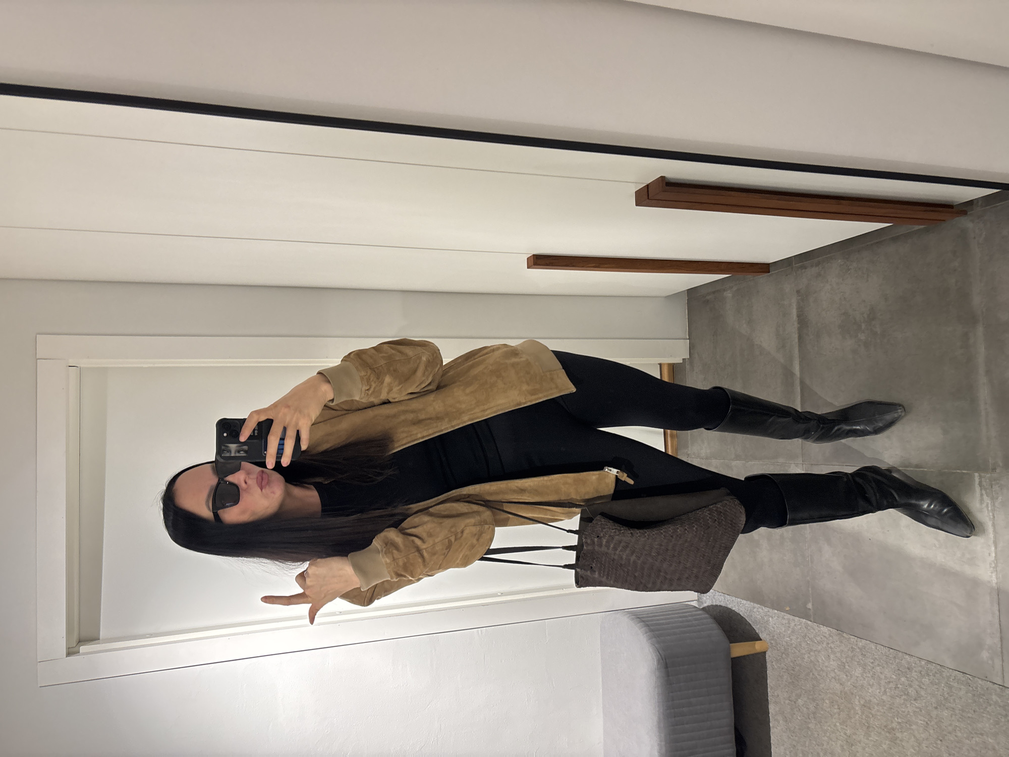 A woman is taking a selfie in a mirror. She is wearing sunglasses, a black top, black leggings, black boots, and a tan jacket. She is holding a cell phone in her right hand and making a hand gesture with her left. A purse is hanging from her shoulder. The background consists of a wall, door, flooring and a grey chair. The location is not evident.