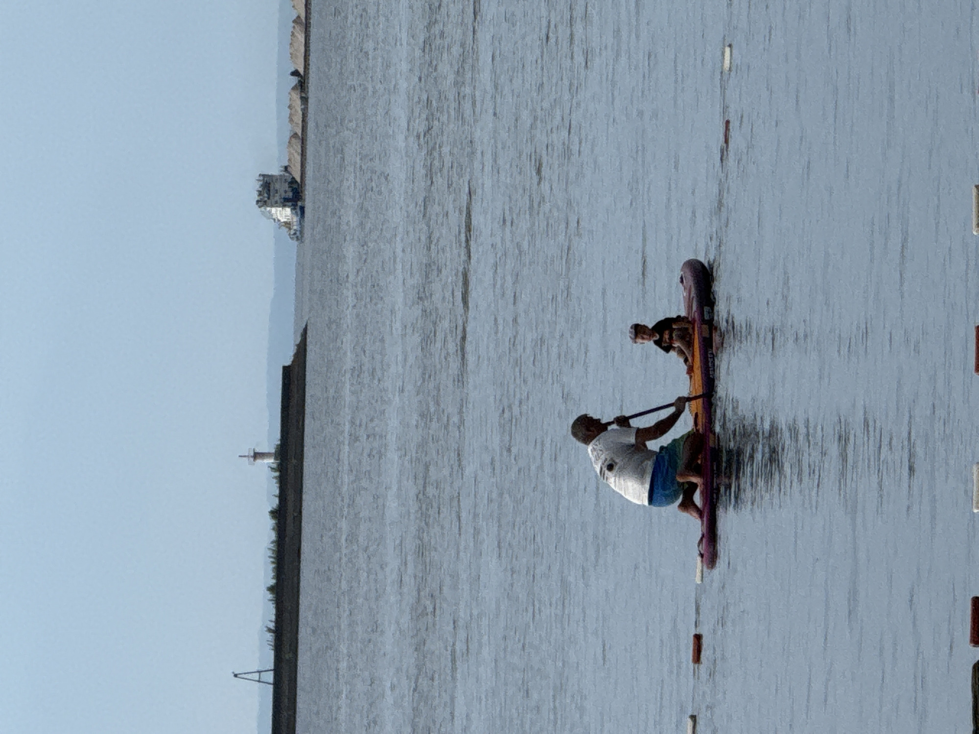In the city of Chaikovsky, Russia, an adult male on a paddleboard is kneeling and holding a paddle, with his arm extended forward. A young child is seated on the front of the same paddleboard, facing forward and wearing a dark t-shirt and shorts. The paddleboard is on a body of water. In the background, a barge with a structure on its deck is visible on the water, and further in the distance, a breakwater with a lighthouse-like structure and a tall, A-frame marker can be seen. The sky is overcast.