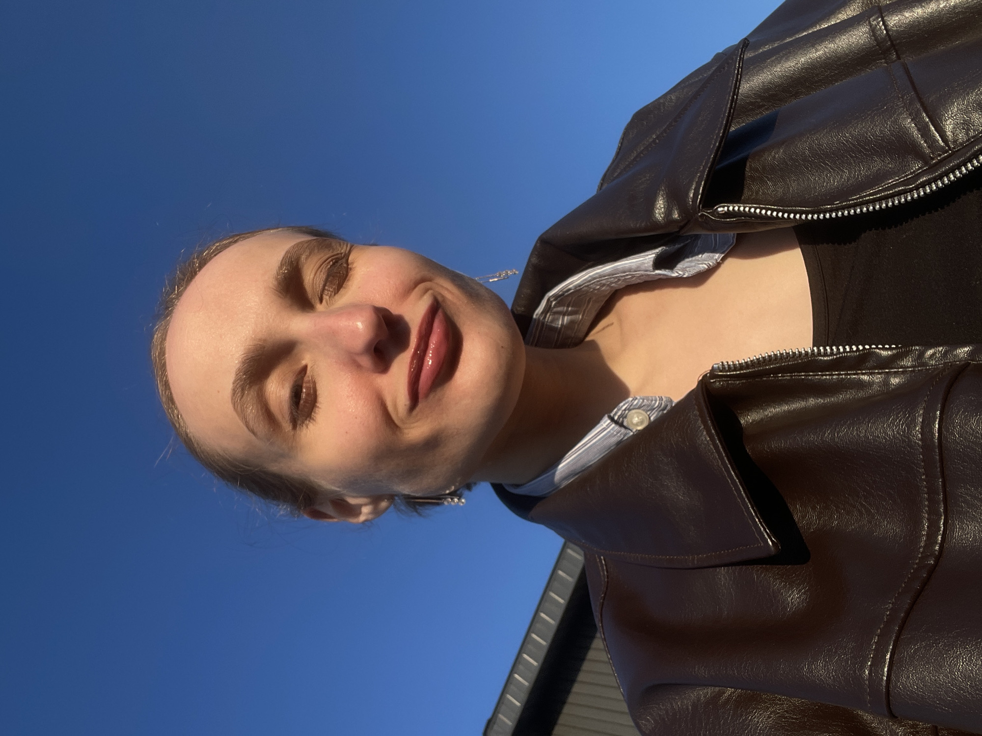 A young woman is captured in a close-up, portrait-style shot, looking slightly upwards towards a clear blue sky. Her eyes are gently closed or squinting, and she has a subtle, serene smile, indicating enjoyment of the bright sunlight. She is wearing a dark brown, possibly faux leather, jacket with a prominent collar and a metal zipper detail. Beneath the jacket, a black top and a light-colored, striped button-down shirt are visible. The dominant background is a vast, cloudless blue sky, suggesting excellent weather and a sunny daytime setting. Warm sunlight casts highlights on her skin, the subtle wisps of her hair, and the textured surface of her jacket. A dark building structure with vertical elements is partially visible in the lower right corner, suggesting an urban or rural outdoor location. No visible text is present. The scene is set in Sovetsko-Nikol'skoe Lesnichestvo, Russia.
