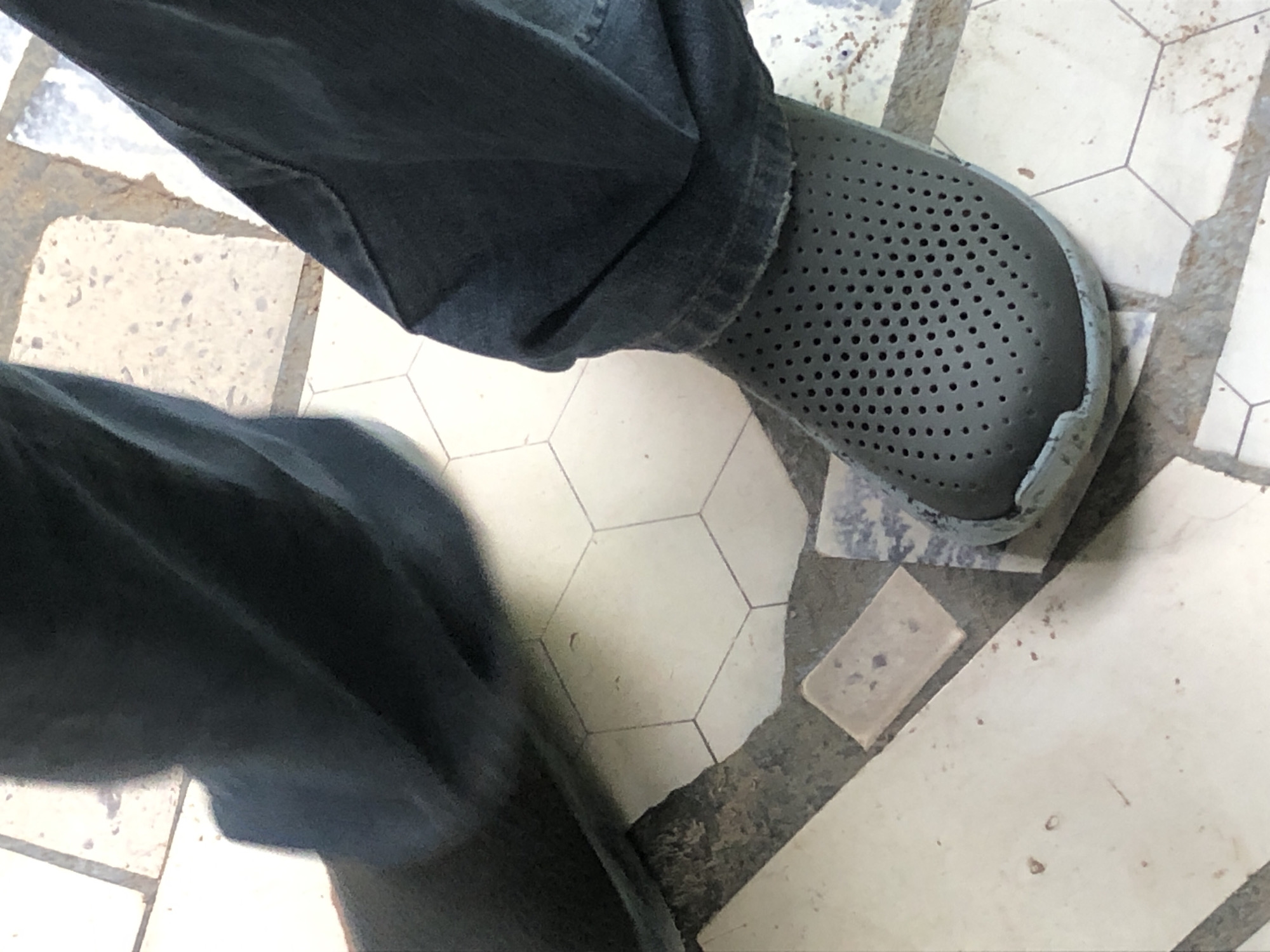 The image shows a close-up view of a person's legs and feet wearing dark blue jeans and dark gray perforated shoes. The person is standing on a floor with hexagonal and rectangular tiles. The tiles are off-white with some darker grout lines. There are also some patches of rough, gray material between the tiles. The lighting suggests an outdoor or well-lit indoor environment.