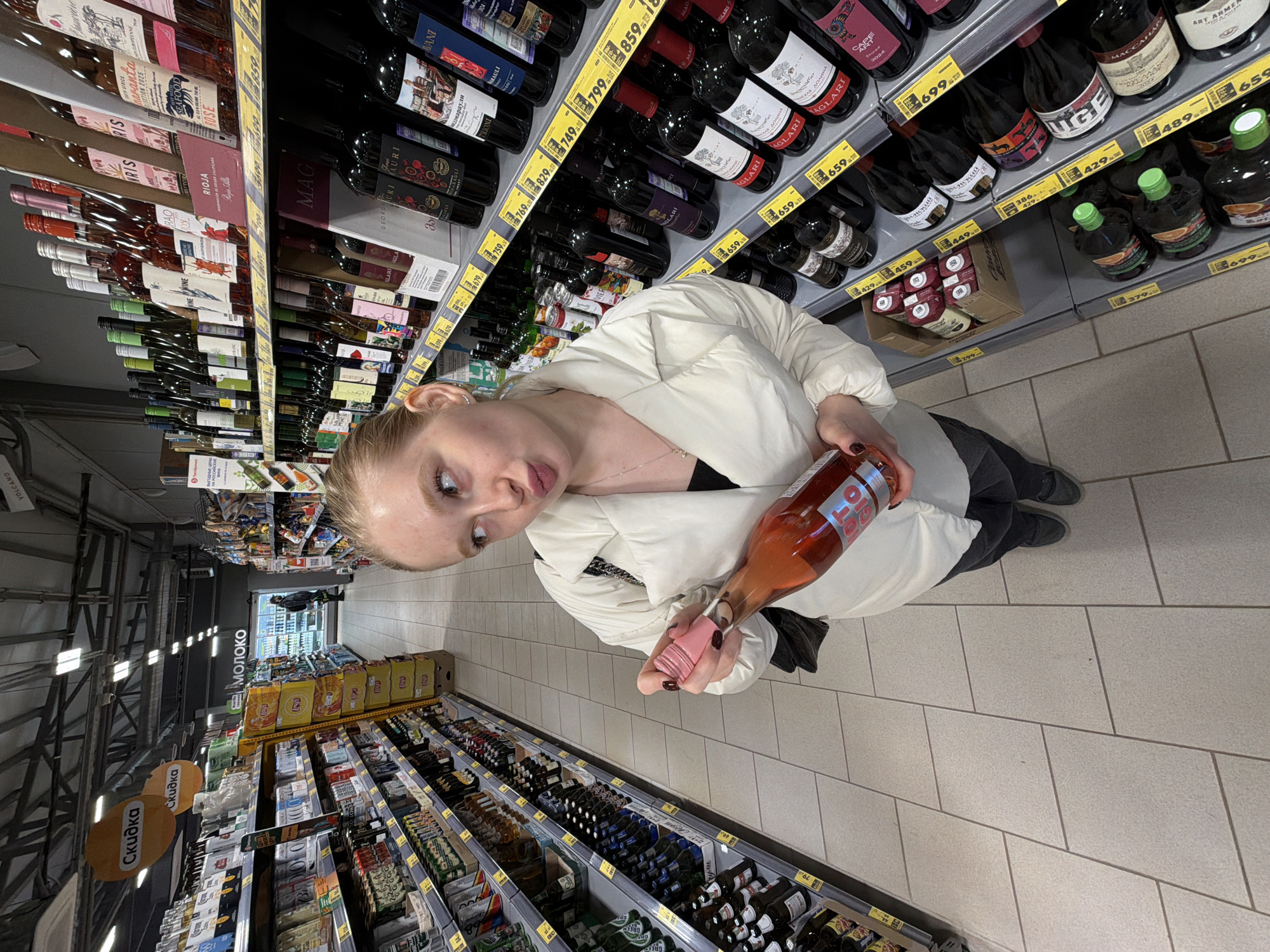 The image depicts a woman holding a bottle of rosé wine in a store aisle. The store appears to be a supermarket or liquor store with shelves stocked with various alcoholic beverages, predominantly wines and beers. She is wearing a white puffy jacket and black pants. In the background, there are shelves filled with wine bottles, each labeled with prices in rubles. Further down the aisle, visible signs indicate the "Moloko" section and promotional "Skidka" signs. The location is likely in Krasnodar, Russia.